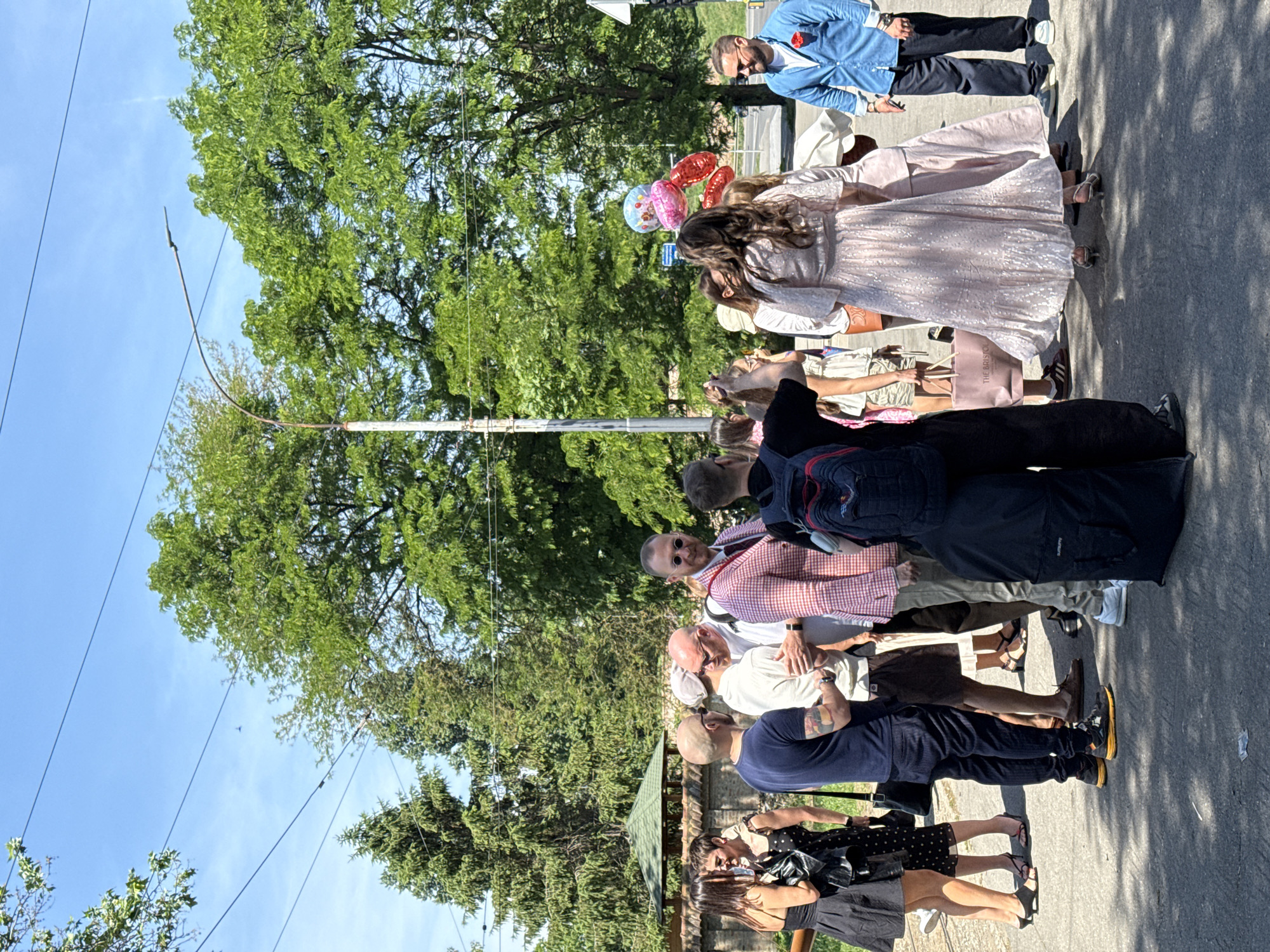 A group of people, including at least one woman in a pale pink dress with balloons, stand along a paved surface, possibly a sidewalk or road, near green trees, and electrical wiring, in an outdoor setting under a bright sky. The location is Belgrade, Serbia. The people seem to be socializing and possibly attending an event or gathering.