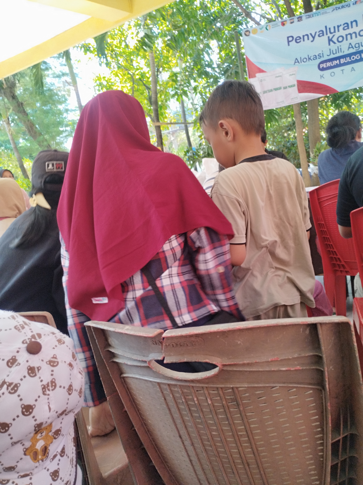 The image shows a group of people sitting in an outdoor setting, likely a public event or gathering. In the foreground, a person wearing a maroon hijab and a plaid shirt is seated, with their back to the camera. To their right, a young boy in a beige t-shirt is also seated. In the background, there is a banner with Indonesian text, which appears to be related to a distribution program. The environment suggests a tropical or subtropical climate, with lush green foliage visible behind the banner. The presence of plastic chairs indicates a functional setup for an event. The location is identified as Surabaya, Indonesia.