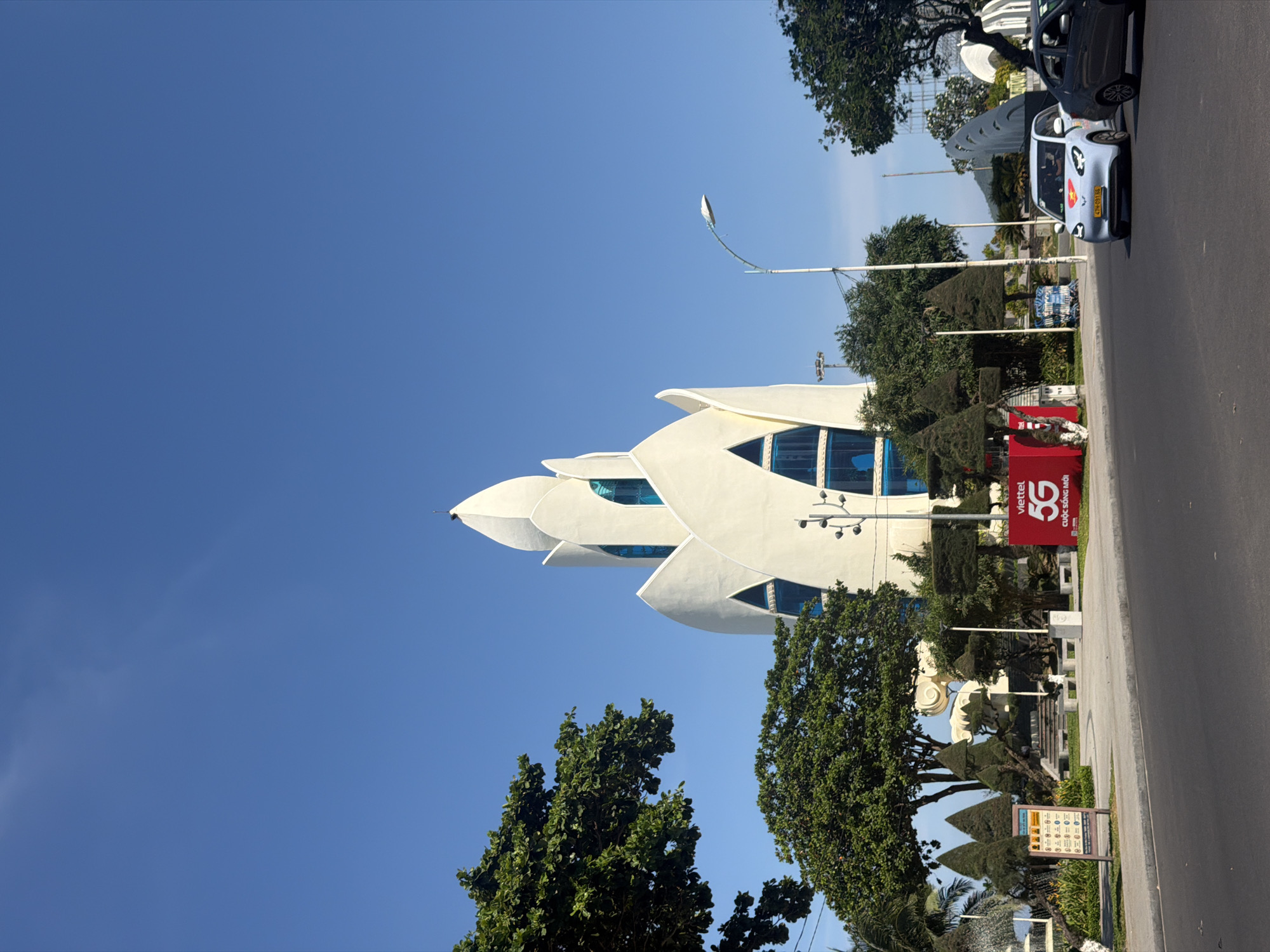 The image shows the main entrance to the Nha Trang Center complex in Nha Trang City, Vietnam, on a clear, sunny day. A distinctive white, lotus-shaped building with blue windows stands prominently in the center of the frame. In the foreground, a paved road with a sidewalk runs horizontally. To the right of the road, a light blue electric car with a Vietnamese flag sticker on its hood is driving, followed by a dark gray sedan.
On the sidewalk, there is a red billboard with "Viettel 5G CUDC SONG MOI" written on it. To the left of the billboard, a bulletin board displays several images and text. Various trees and manicured bushes are scattered throughout the scene, with a large, leafy tree dominating the left side of the image. In the background, more trees and some partially visible structures can be seen. The sky is a vibrant, clear blue.