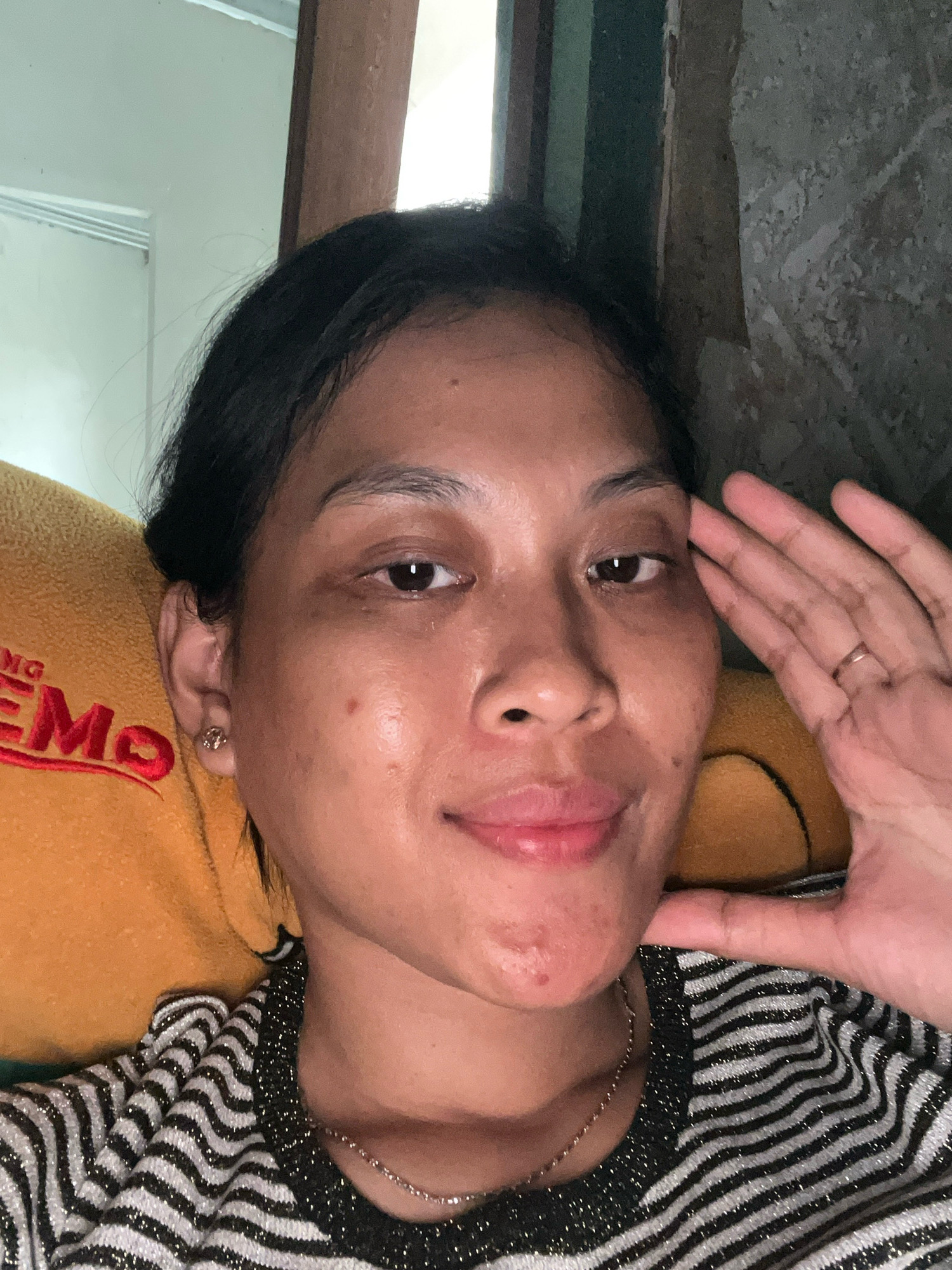 A young woman with dark hair is taking a selfie. She is leaning against a yellow pillow with red lettering. She is wearing a striped shirt and a necklace. Her right hand is raised to her face. The background appears to be an indoor setting with a wall and a window. The image seems to have been taken in Kecamatan Pati, Indonesia.