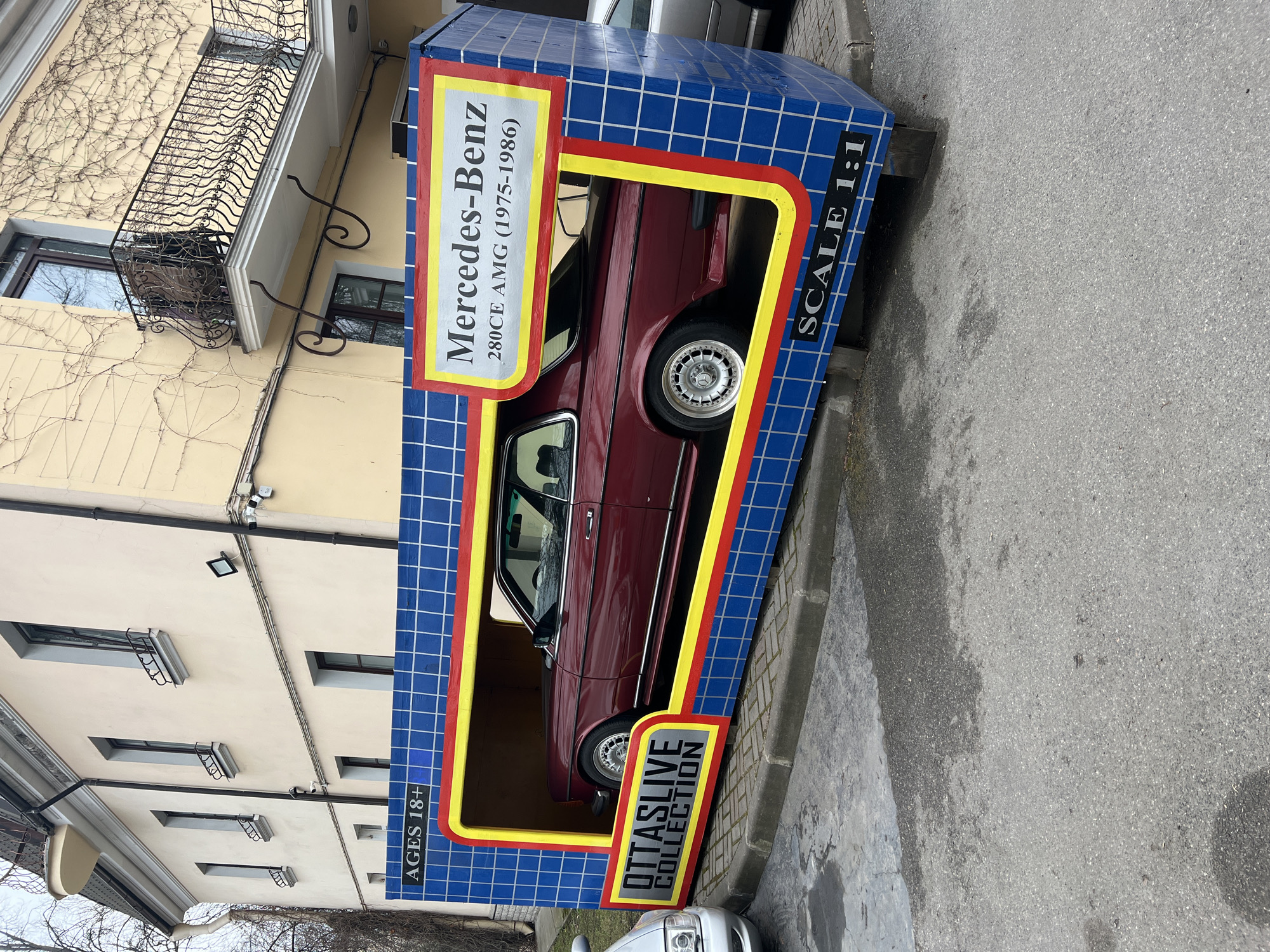 The image captures a life-size model of a Mercedes-Benz 280CE AMG (1975-1986) seemingly encased in a toy box. The box features a blue grid pattern, with a yellow and red trim around the "window" displaying the car. Text on the box includes "Mercedes-Benz," "280CE AMG (1975-1986)," "OTTASLIVE COLLECTION," "AGES 18+," and "SCALE 1:1." The car is maroon in color, visible through the cut-out section of the box.
The scene is outdoors, with the model placed on a pavement or street next to a building. The building has a light yellow facade with multiple windows, some featuring small railings. There is a balcony with ornate black metalwork on the upper part of the building. Bare vines crawl on the side of the building. The road surface is grey.
Based on the "OTTASLIVE COLLECTION" branding and available information, the location is Saint Petersburg, Russia.