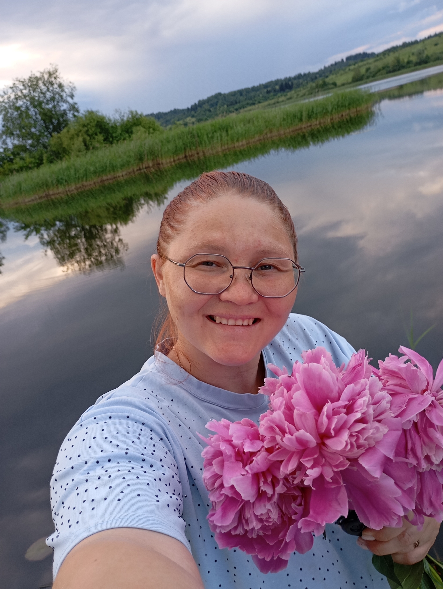 A woman with auburn hair and glasses is taking a selfie outdoors. She is wearing a light blue t-shirt with dark polka dots. In her hands, she is holding a bouquet of vibrant pink peonies. Behind her, a calm body of water reflects the sky and a treed shoreline. The sky is partly cloudy with a mix of grey and white clouds. The overall scene suggests a natural setting, possibly near a lake or river. The location is identified as Ryabki, Russia.