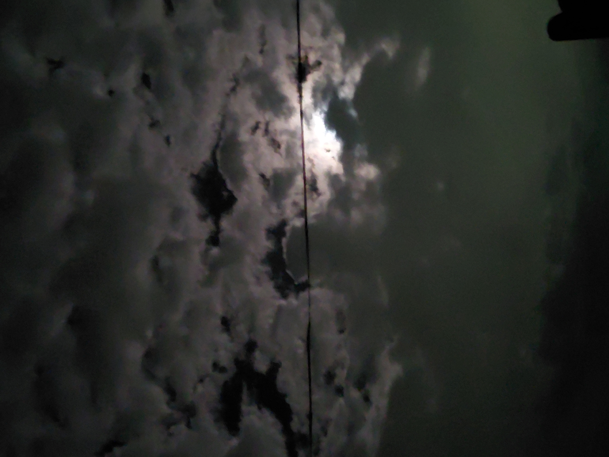 The image displays a dark, cloudy night sky, likely illuminated by the moon, though the moon itself is mostly obscured. Dense, dark grey and black clouds dominate the frame, with a bright, diffused light source breaking through a gap in the clouds towards the upper-center, casting a luminous glow. This light highlights the edges and textures of the surrounding clouds.
A thin, dark vertical line, possibly a utility wire or cable, runs down the center of the image, bisecting the bright light and clouds. To the far right, a dark, indistinct shape, potentially a tree or part of a structure, is silhouetted against the slightly lighter background of the sky. The overall color scheme is predominantly dark, with shades of black, charcoal grey, and hints of a deep greenish-blue in the sky, contrasting with the bright white and light grey of the illuminated clouds. The image captures a serene yet somewhat eerie nocturnal scene. No specific activities or visible text are present. The setting is outdoors at night, in Santa Ana, Venezuela.