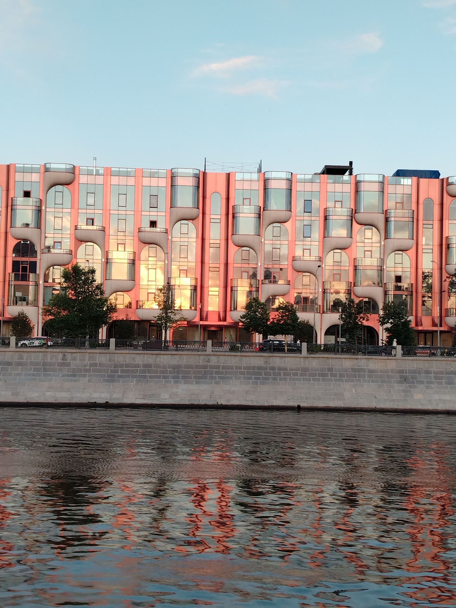 This is a photograph of a modern, pink building with large glass windows, reflecting the sunset, situated on the embankment of a river in Moscow, Russia. The building has a distinctive architectural style with curved balconies and repeated rectangular window shapes. Trees line the area between the building and the river. The river in the foreground reflects the colors of the sunset and the building. The sky above is a light blue with a few wispy clouds. The embankment is made of stone. There are no people visible in the image, but there are a few cars parked along the sidewalk in front of the building.
