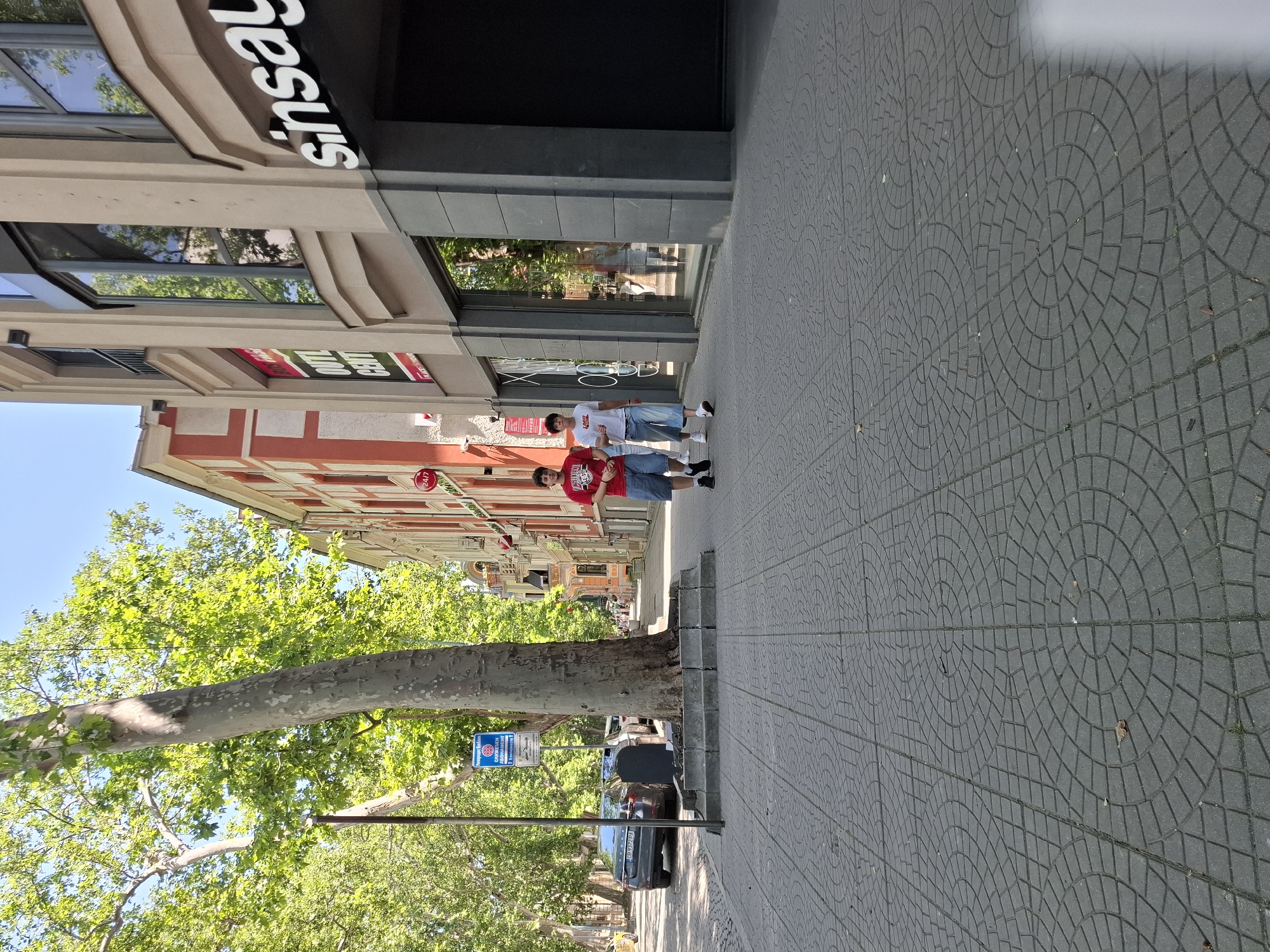Two young men walk along a paved sidewalk in a city. The man on the left wears a red t-shirt and blue shorts, while the man on the right wears a white t-shirt and light blue capri pants. They are walking away from the camera, towards a building on the right side of the image. The building has large windows and a prominent "Sinsay" sign. To the left of the sidewalk, a large tree with green foliage is visible, and further down the street, cars are parked along the road. A street sign with Bulgarian text is mounted on a pole near the tree. The sky is clear and blue. The sidewalk is made of grey interlocking pavers arranged in a circular pattern.