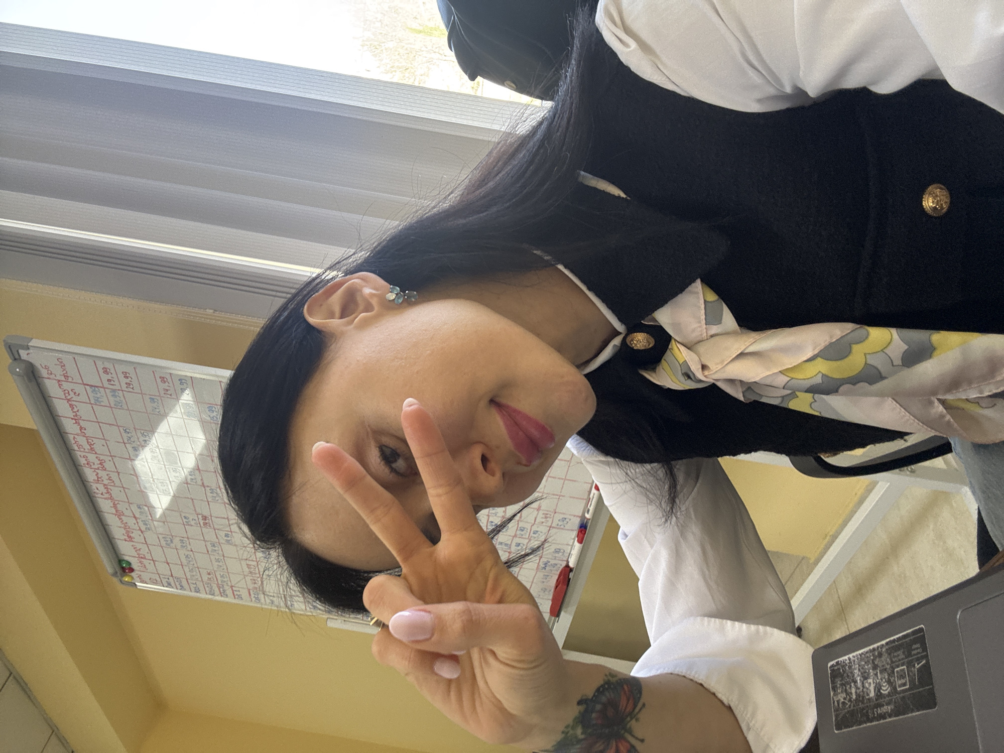 A woman is captured in a selfie, holding her right hand in a peace sign and playfully sticking out her tongue, indicating a friendly and lighthearted mood. The image is rotated, showing her head tilted to the right. She has long, dark hair and is wearing a white long-sleeved shirt, a black sleeveless vest adorned with gold buttons, and a patterned scarf featuring cream, yellow, and grey hues around her neck. She wears small, light-colored earrings and has light pink nail polish. A tattoo of a butterfly is visible on her left wrist.
The setting appears to be an indoor environment, likely an office or classroom, based on the presence of a whiteboard behind her. The whiteboard displays handwritten text and numbers in both red and black markers, including multiple instances of prices like "19.99", "24.99", and "39.99". Significantly, some text on the whiteboard is written in Georgian script, confirming the location context of Tbilisi, Georgia.
Natural light suggests it is daytime, filtering in from a window partially visible through horizontal blinds in the upper background. The walls are a light yellow or cream color. Part of a grey laptop, featuring a sticker with small graphics and text including "SUS Advisory", is visible in the lower right foreground. A black office chair is partially seen behind the woman.