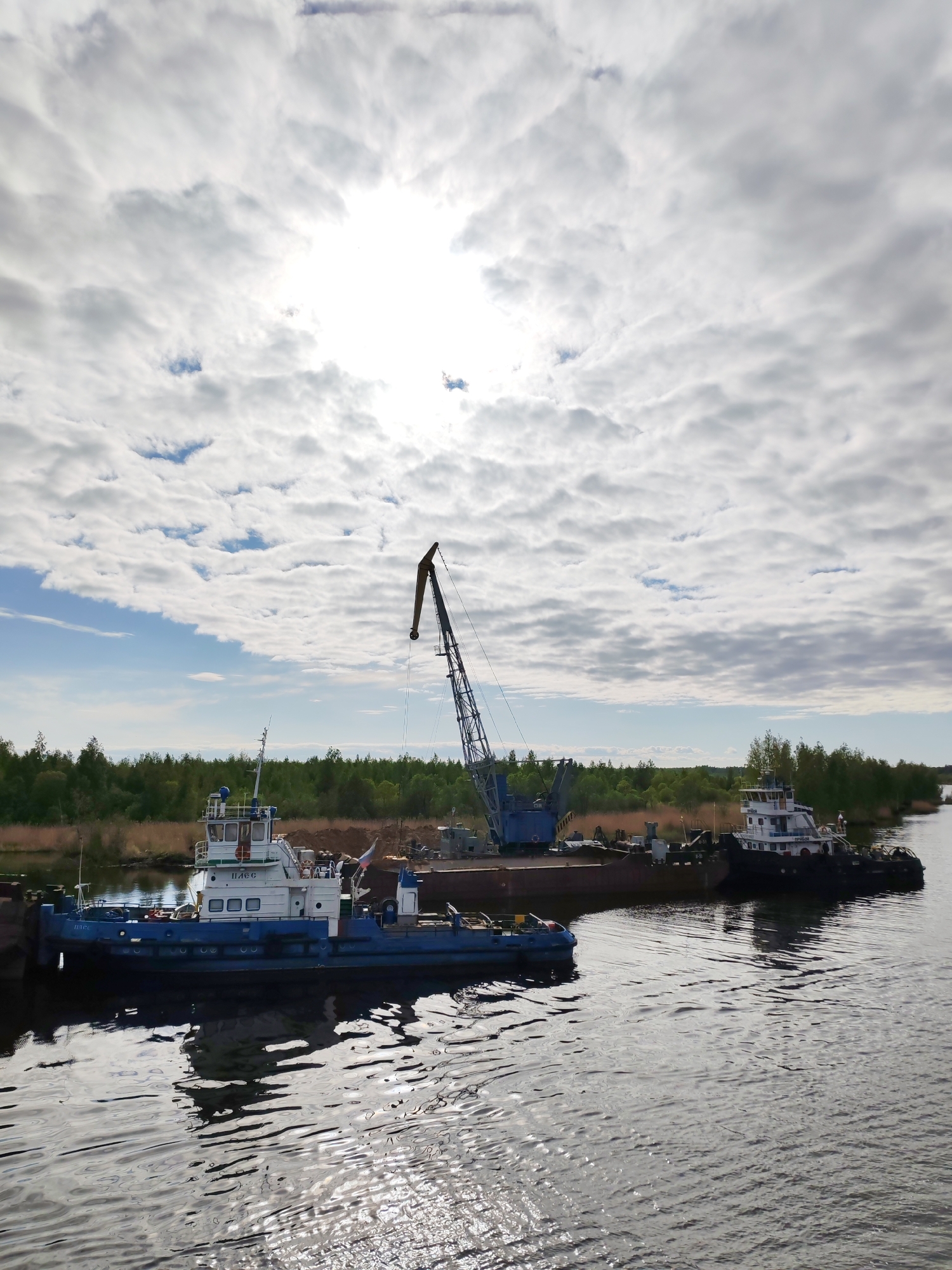 The scene depicts a waterway in Ozernoe Ust'e, Russia, with two tugboats and a barge equipped with a crane. The tugboats are positioned on either side of the barge, appearing to be moored. The crane is centrally located on the barge, with its boom extending upwards. The waterway's surface reflects the sky and vessels. The shoreline is visible in the background, lined with trees and vegetation. The sky is overcast with clouds, obscuring the sun.