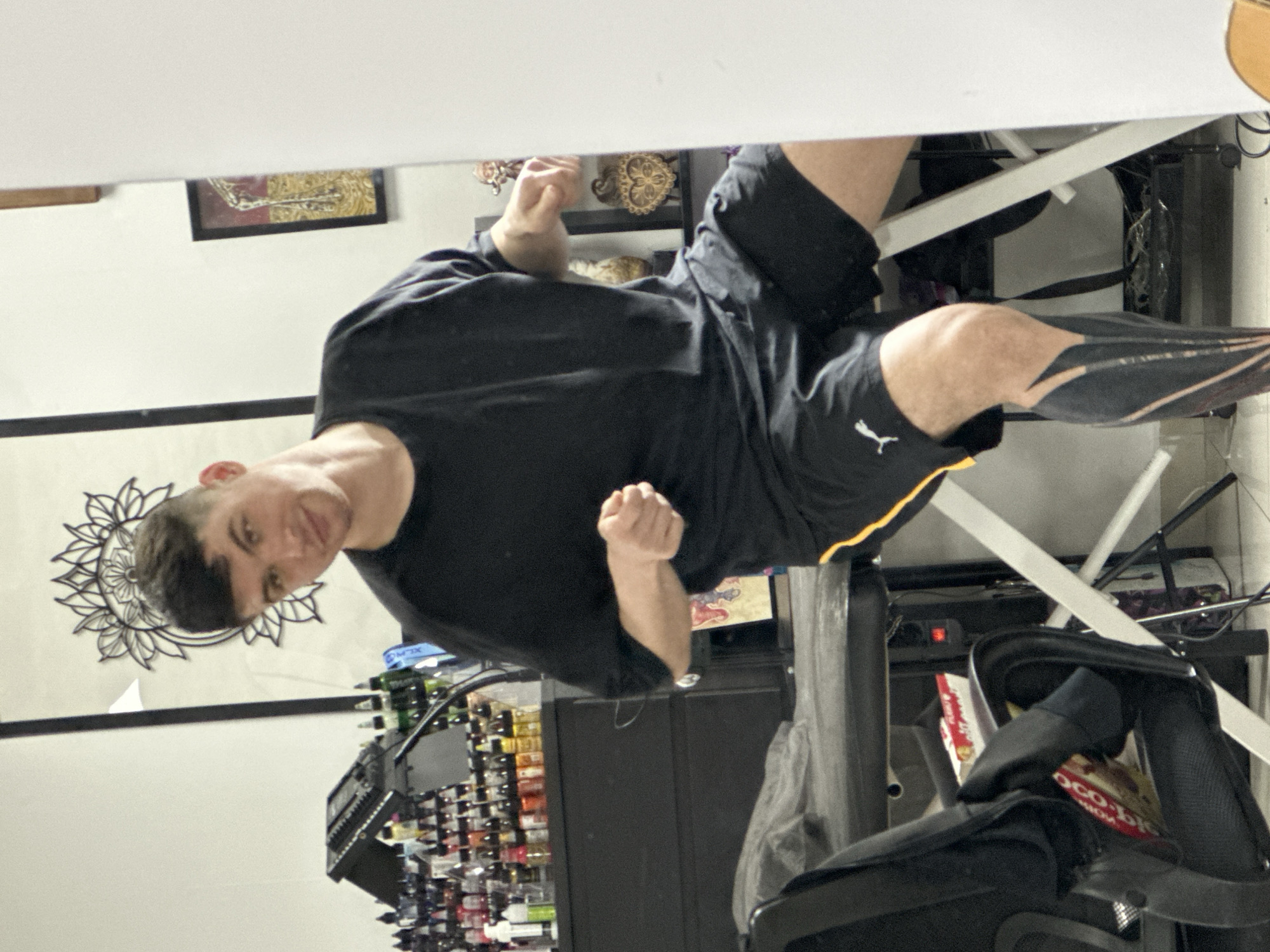 The image shows a man in a black t-shirt and black shorts with a yellow stripe, seated at a desk. He has short, dark hair and is looking at the camera with his fists clenched. Behind him is a wall with a mandala design and a shelf with numerous bottles, possibly tattoo inks. A black desk chair is visible in the foreground. The location of the scene is unknown, with no definitive indicators suggesting Rostov-on-Don, Russia.