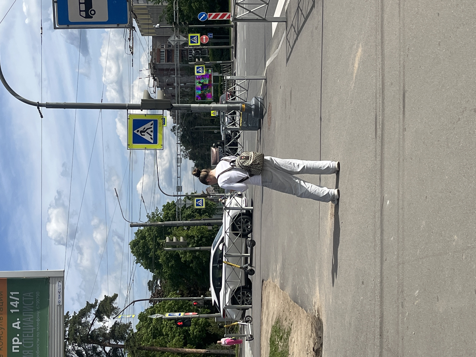 A bright daytime street scene in an urban environment, likely Saint Petersburg, Russia, features a woman standing on the edge of a paved area next to a road. The woman, seen from the side, has her hair in a bun, wears a white long-sleeved top, light grey pants, and a small backpack with a distinctive brown and beige pattern, possibly from the brand Gucci. Her shadow is cast long on the ground, indicating bright sunlight.
To her left, a multi-lane asphalt road stretches into the distance, with a white car and two electric scooters parked alongside it. A red traffic light is visible further down the street, and a pedestrian crossing sign (blue square with a white pedestrian icon) is prominently displayed on a pole above her. Other road signs, including a blue bus stop sign, a triangular yield sign, and a red circle 'no entry' sign, are also visible. Metal barriers line the sidewalk, separating it from the roadway in some sections.
In the background, various buildings with green trees interspersed create an urban canopy under a partly cloudy sky. To the far left, a billboard displays text in Russian, including "пр. д. 14/1", "НЯ СПЕЦИАЛИСТА", and contact information "по телефону: 6790790", along with "ID 104". The road surface shows signs of wear, with some cracks and lighter-colored streaks. The overall impression is a bustling street on a clear, sunny day.