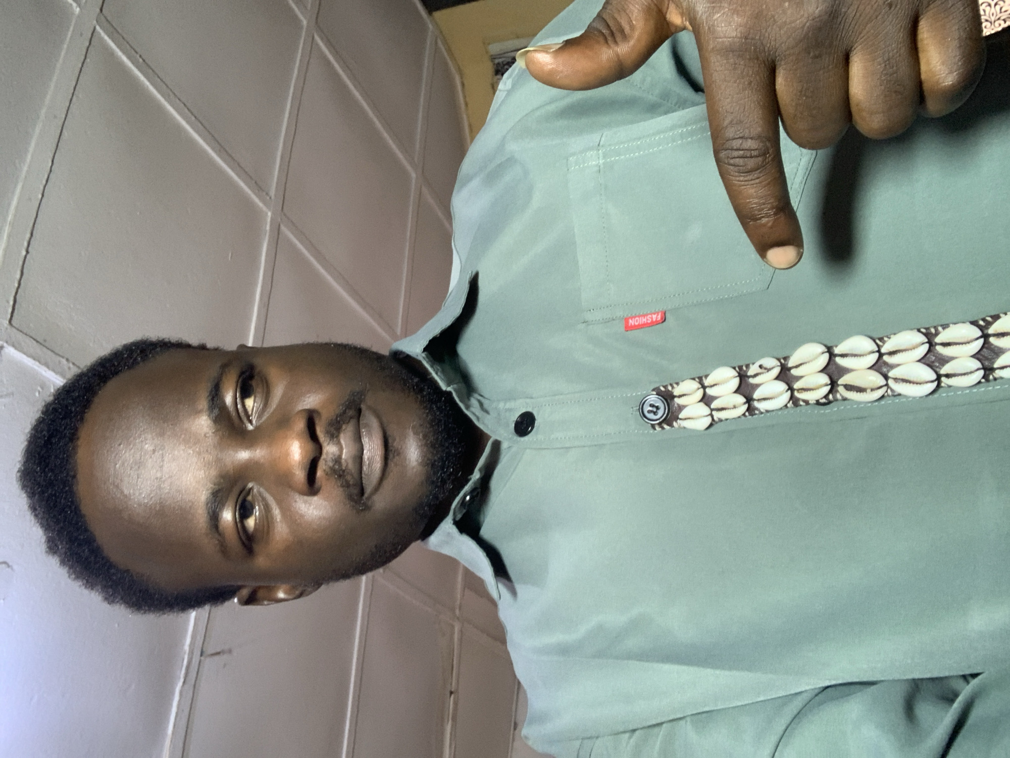 A man, appearing to be of African descent with dark skin and a goatee, is captured in a medium shot, looking to his right with a neutral expression. He is wearing a light green collared shirt adorned with a decorative strip of small, light-colored shells running down the placket. A small red tag with the word "FASHION" is visible on the left breast pocket of the shirt. His right hand is raised, with his thumb and index finger extended in a pointing gesture towards the viewer. The background consists of a white tiled ceiling. The overall lighting suggests an indoor setting. The image does not provide any specific information about the location within Jos, Nigeria.