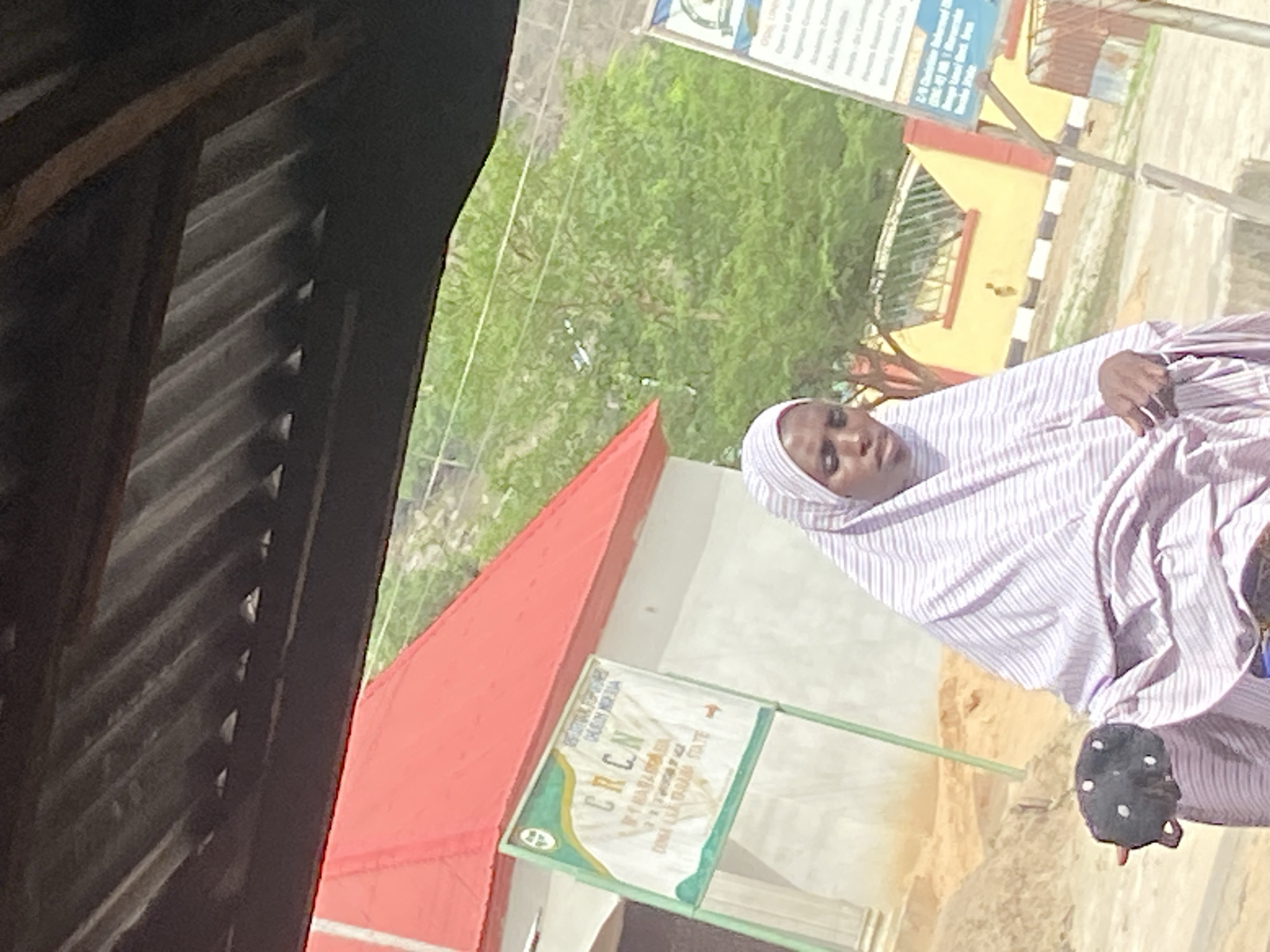 An outdoor, daytime image depicts a woman standing near buildings and signboards in what appears to be Mararaba, Nigeria. The image is oriented vertically but should be viewed as if rotated counter-clockwise to be upright.
In the center-right, a woman is standing, facing slightly to her left. She is wearing a light-colored, possibly white or light purple, vertically striped head covering and a matching or similarly striped garment that covers her body. Her expression is neutral. She is wearing dark sandals with white circular embellishments on her right foot.
The immediate left foreground shows a dark, possibly wooden or corrugated metal structure, perhaps part of a veranda or shelter. Beyond this, a building with a prominent red roof and light-colored walls is visible. In front of this building, a green and white sign is clearly visible. The most legible text on this sign is "ERCN" at the top, with a stylized cross symbol to its left. Below "ERCN" is "P.H. BAHAIMASU". Other, smaller text is present but difficult to decipher.
Further in the background, a yellowish building with black and white striped markings on a low wall or curb is visible. Above this building, another blue and white signboard is partially visible, containing blurry text where "CPCN" and "Nigerian Council of..." can be faintly discerned. Green foliage and trees are visible throughout the background, suggesting a natural environment alongside the urban structures. The ground appears to be a mix of dirt and sand. The overall lighting suggests it is bright daylight.