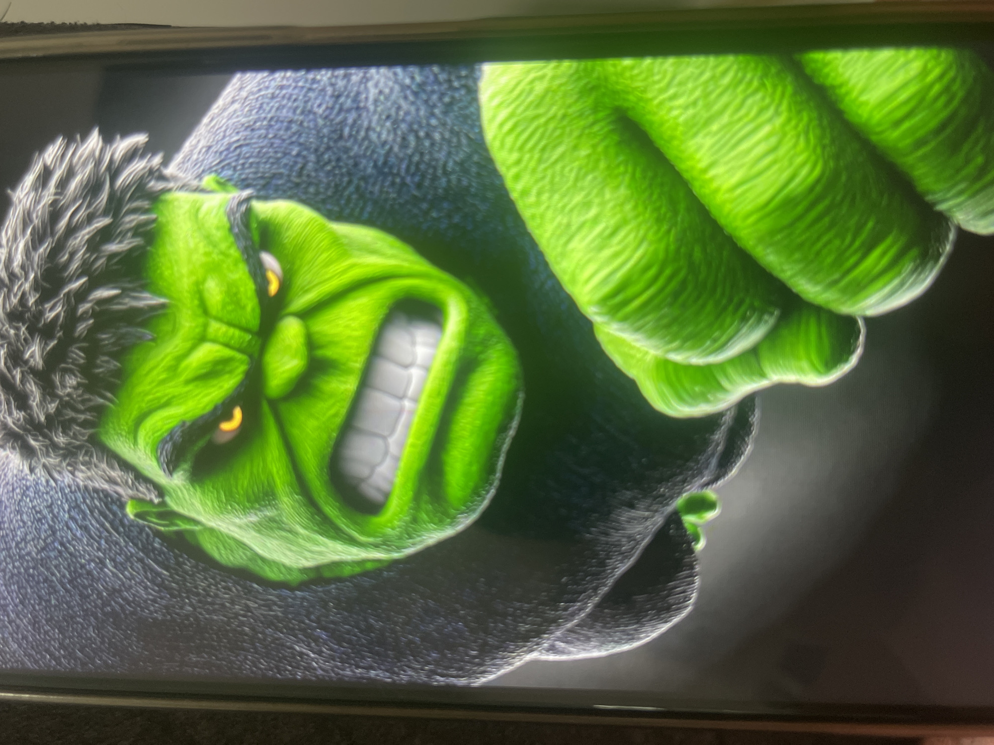 The image is a close-up, low-angle shot of a depiction of the Hulk. The character is rendered in vivid green with yellow eyes, a snarling expression, and coarse gray hair. He is shown from the chest up, with his right hand prominently in the foreground, appearing to be reaching out or about to strike. His body is covered in a dark, textured material, possibly representing his clothing or skin. The background is dark and out of focus, emphasizing the Hulk's presence. The image appears to be a digital artwork or a screenshot from a screen, as evidenced by the slight glare and border on the right side. There is no discernible information or context within the image that would suggest a specific location in Bishkek City, Kyrgyzstan.