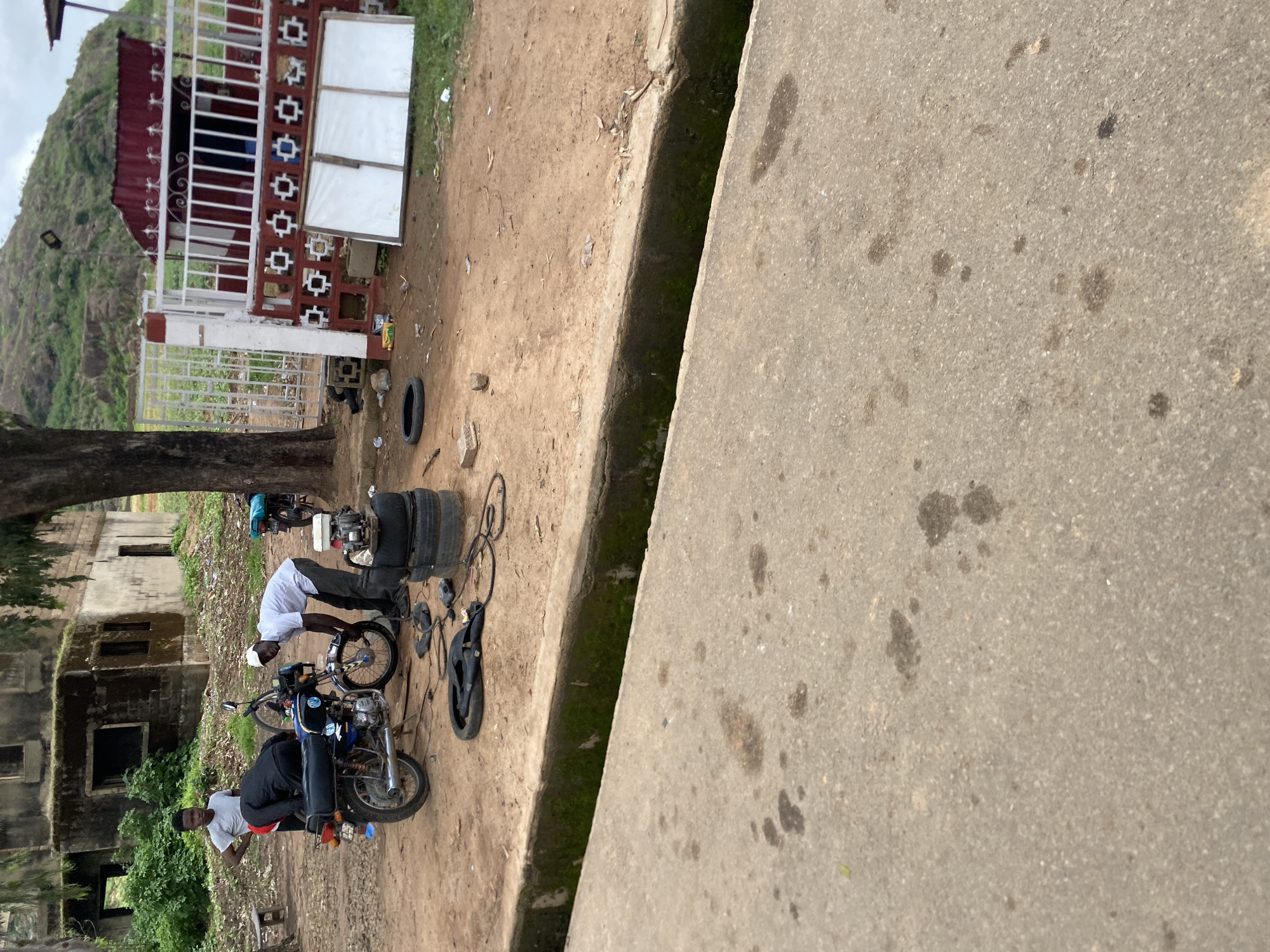 Two young men are working on a motorcycle in a dirt lot. The motorcycle is blue and black. One man, wearing a white shirt and a white cap, is bent over the front wheel of the motorcycle, appearing to be working on it. He is wearing grey pants. The second man, wearing a grey t-shirt and dark pants, stands behind the motorcycle, looking towards the first man. Another motorcycle, blue and black, is parked behind them. In the background, there is a dilapidated, multi-story building with moss growing on its exterior. A large tree is to the left of the building. To the right of the building, there is a red metal structure with white fencing. The ground is covered in dirt and scattered with various objects, including tires, tools, and debris. The scene is located in the city of Jos, Nigeria.