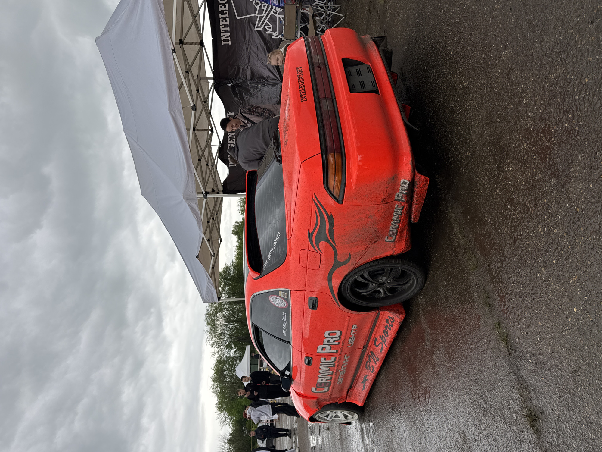 The image shows a red sports car with the logos "CERAMIC PRO" and "BN Sports", parked on a wet surface. The car appears dirty, suggesting it may have recently been used in a race. The car is parked beneath a tent with the word "INTELEGENT" printed on the side. Several people are standing around in the background. The license plate says “INTELEGENTOFF” and the user’s Instagram handle is printed on the window. The image was likely taken at a car event in Pavlovskaya, Russia, based on the Cyrillic text "ДЕТЕЙПИНГ ЦЕНТР" (Detailing Center) on the car.