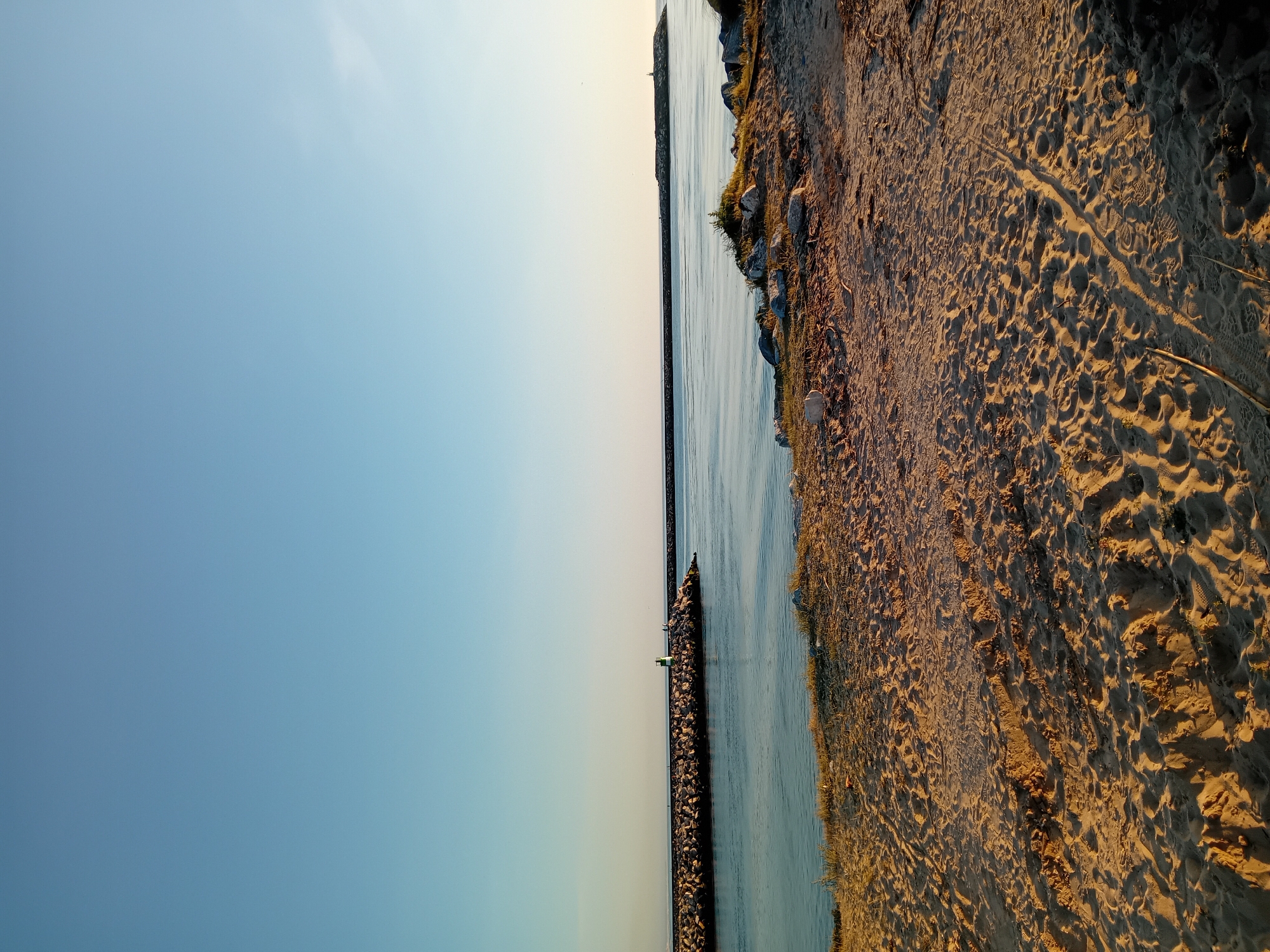 The image shows a beach scene in Figueira da Foz, Portugal. The composition is divided into thirds horizontally. The left third is a clear, blue sky transitioning to a pale yellow near the horizon. The middle third contains a narrow channel of water, bordered by stone breakwaters on each side. The right third consists of a sandy beach marked with numerous footprints. The sand is illuminated by a warm light, likely from the setting sun, casting shadows and highlighting the textures of the sand. Vegetation is sparsely distributed along the edge of the beach. The image suggests a tranquil, seaside environment.