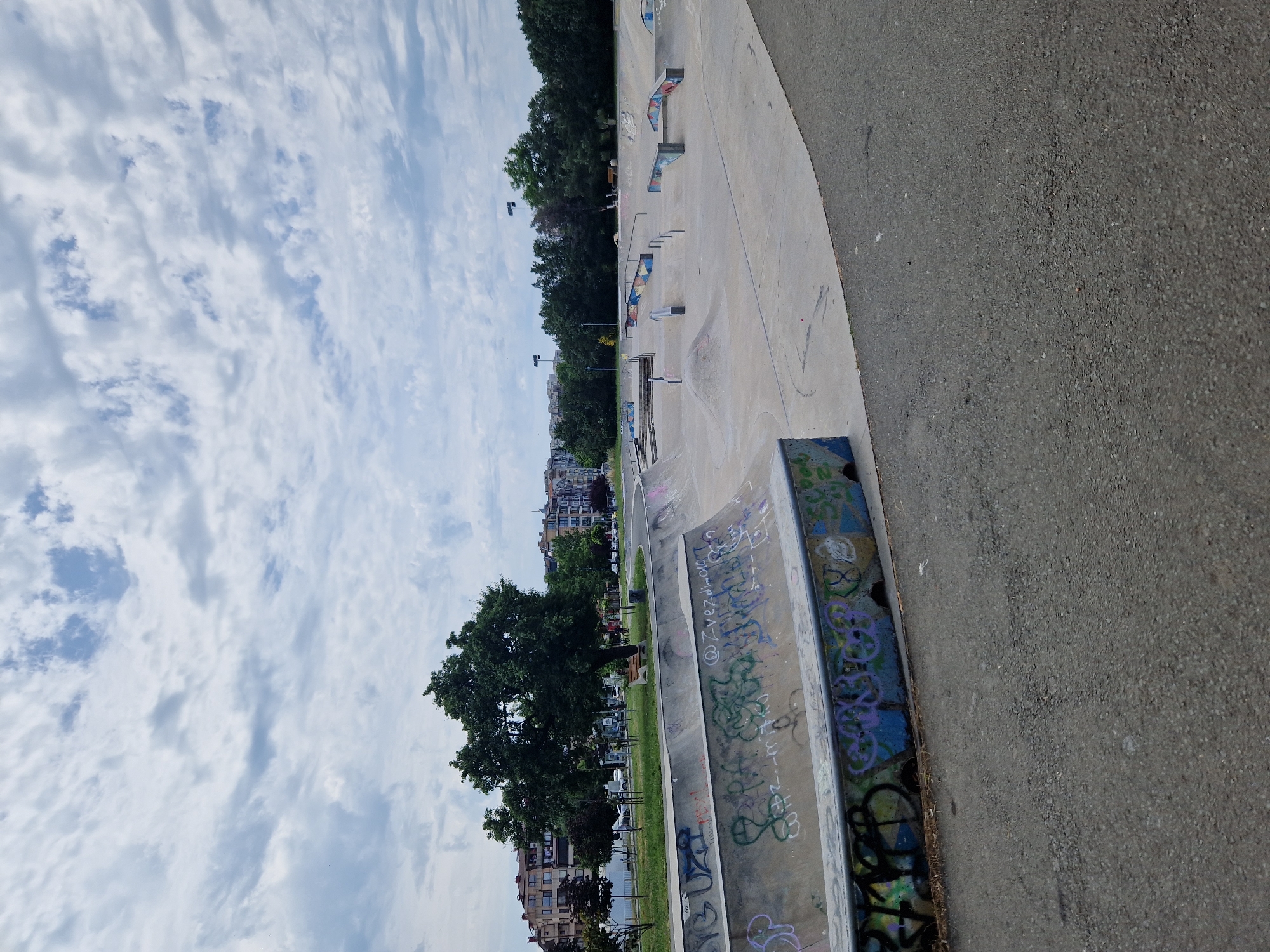 A public skatepark with concrete ramps and rails is depicted under a cloudy sky. The foreground features a section of the skatepark with graffiti on its walls. Beyond the skatepark, there is a tree-lined area with buildings and parked cars. The overall scene suggests an urban recreational space on a partly cloudy day.
