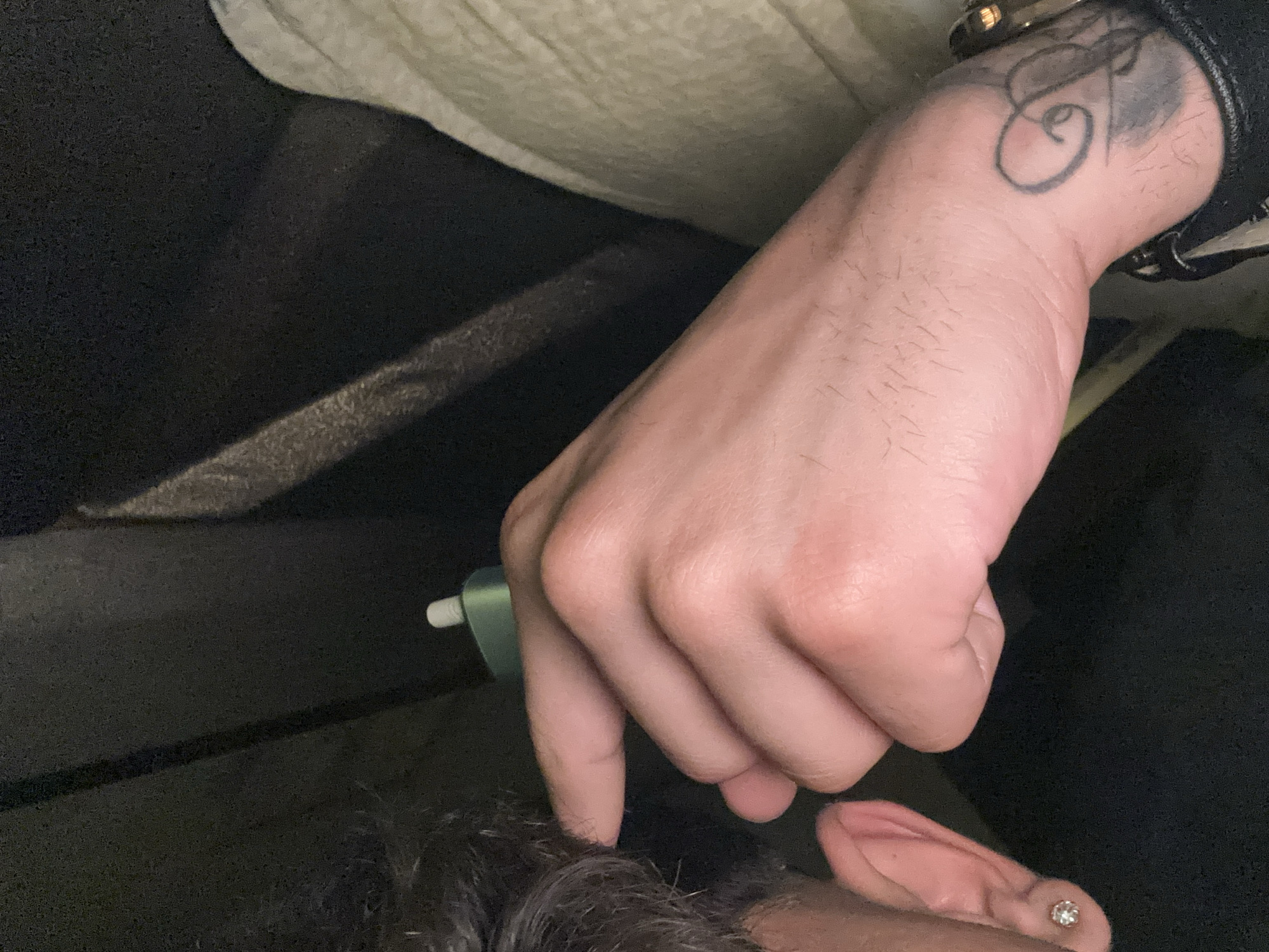 The image displays a close-up, dimly lit view of an individual from a high angle. On the left, the side of a head is visible, featuring short, dark, textured hair and an ear adorned with a small, round, clear stud earring. A hand with pale skin and fine hair on the forearm is centrally positioned, holding a compact, light green electronic device with a white tip. The index finger of this hand is extended towards the head, near the ear, suggesting an interaction with that area. On the wrist, a dark, abstract tattoo with swirling lines is visible, partially obscured by a watch with a gold-toned casing and a black strap. The background is dark and indistinct, showing hints of dark, possibly upholstered fabric on the left and a lighter, textured fabric, potentially cream or off-white, on the right, indicating an indoor setting with subdued lighting.