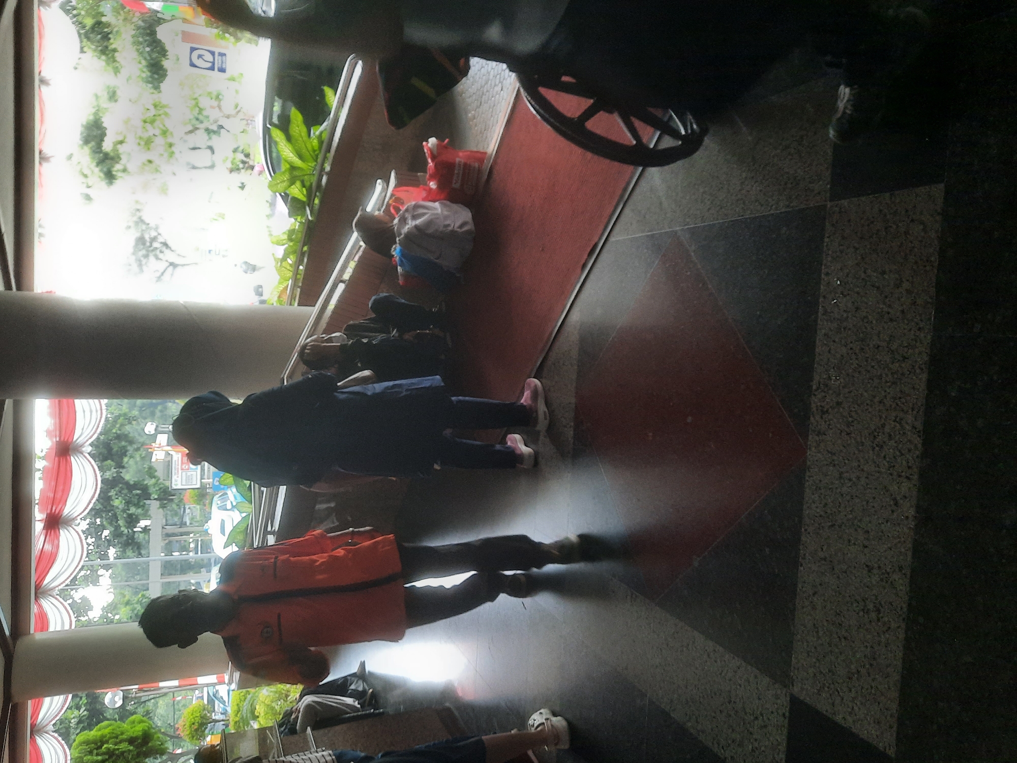 The image depicts an indoor scene with several individuals and various objects. In the foreground, a person is walking away from the camera, wearing an orange shirt and dark pants. To their right, another person in a dark blue outfit and headscarf is also walking. Further back, several people are seated on a red rug, some with bags and luggage. One individual is partially obscured by a wheelchair in the far right of the frame. The environment appears to be a public space with a patterned floor and large windows or open doorways revealing greenery and trees outside. The light suggests it is daytime. The location is specified as West Jakarta, Indonesia.
