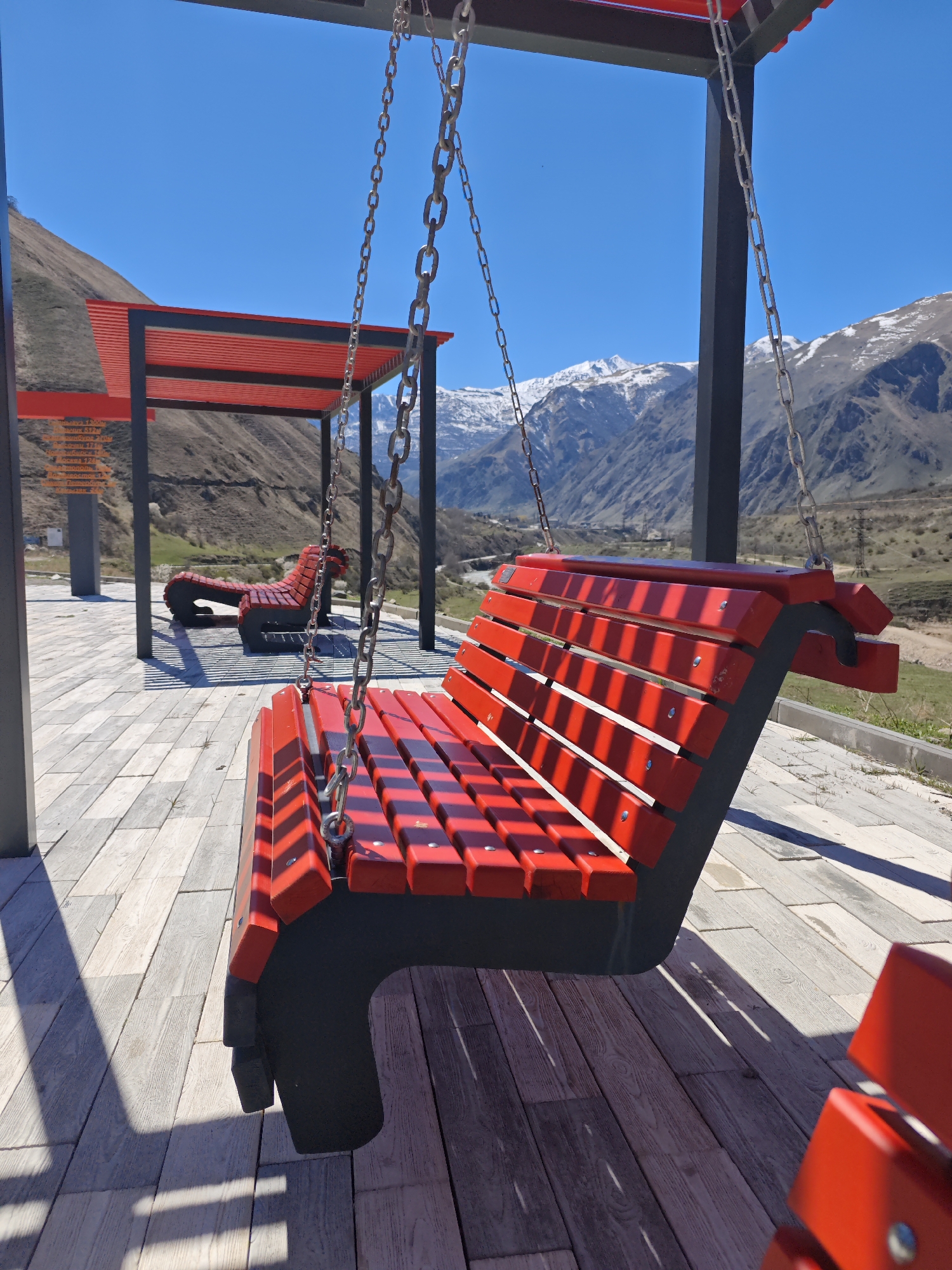 The image captures a scenic view in Tyrnyauz, Russia, featuring a modern seating area with red-and-black benches. The primary subject is a suspended bench, hanging by metal chains, displaying red wooden slats and black structural supports. In the background, another stationary bench mirrors the color scheme. Overhead, red-and-black shelters provide shade. The ground is covered in light gray paving stones, creating a geometric pattern. Beyond the seating area, rolling hills and snow-capped mountains form a striking backdrop under a clear blue sky. The environment appears to be a well-maintained park or public space.