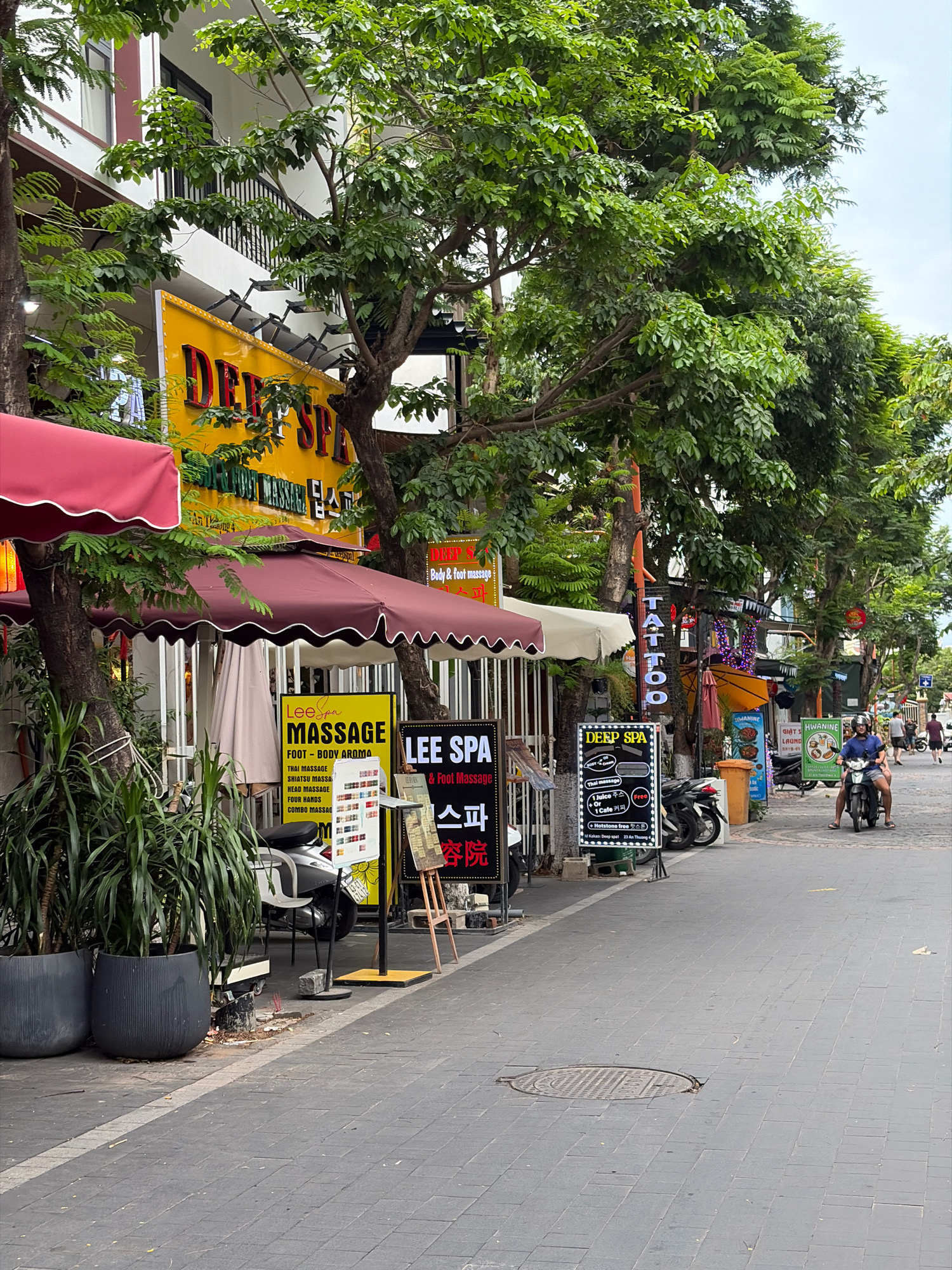 The image shows a street scene in the city of Đà Nẵng, Vietnam, likely a commercial area with several businesses catering to tourists. The foreground features a sidewalk paved with dark gray rectangular tiles, with a white line marking the edge. To the left, two large dark gray planters contain tall, leafy green plants.
Several businesses are visible along the street. A prominent yellow sign with red lettering reads "DEEP SPA", with other smaller signs advertising "MASSAGE", "FOOT - BODY AROMA", "LEE SPA", and "TATTOO". Some of these signs are written in English and what appears to be Korean script. There are red and burgundy awnings providing shade over outdoor seating areas for some of the establishments.
In the middle ground, a silver scooter is parked on the sidewalk. Further down the street, a man wearing a dark blue shirt and shorts is riding a motorcycle with a helmet. Other people are walking in the distance.
The background is dominated by a building with white and dark trim, and lush green trees with dense foliage line the street, casting shadows. The sky is overcast. The overall impression is of a bustling street with various services and shops, typical of a tourist district.