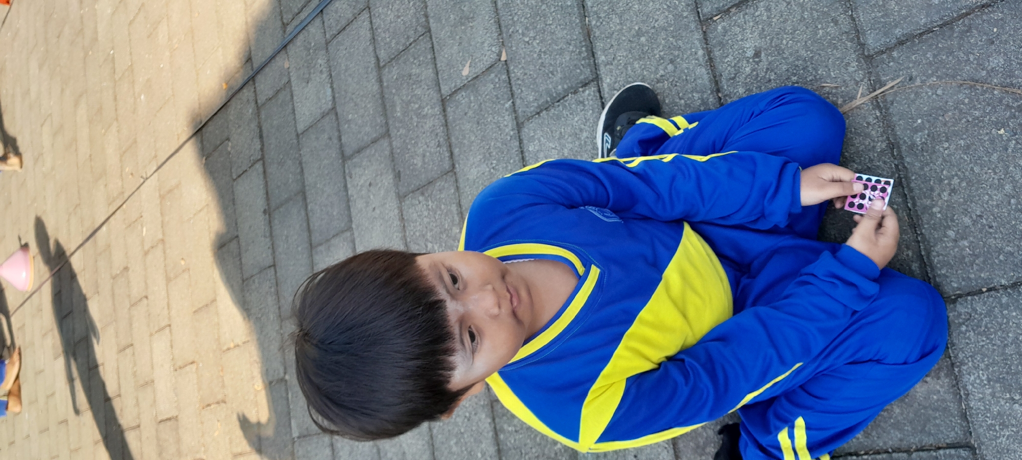 A young boy with dark hair is sitting on a paved surface, possibly a sidewalk or courtyard, in Rogojampi, Indonesia. He is wearing a blue and yellow tracksuit. His hands are holding a small, square object with a pink and white pattern and black circles, resembling a sticker or a game piece. The boy appears to be looking down at the object. The ground is made of interlocking gray pavers, and shadows are cast from an unseen light source, suggesting it is daytime. A thin dark line runs across the paved surface, possibly a wire or a crack. In the blurred background, a pink cone and shadows of people are visible.