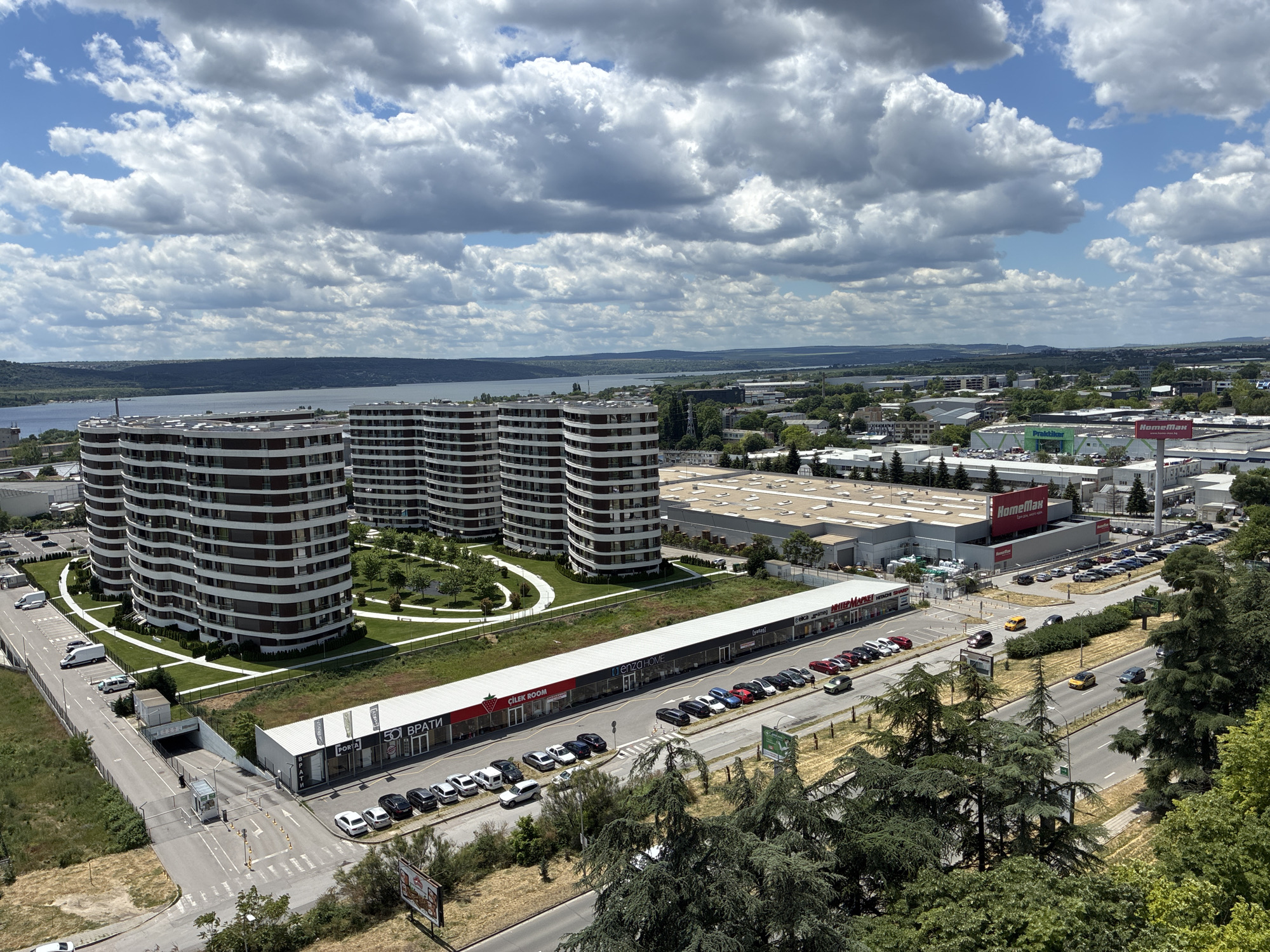 The image depicts a high-angle view of an urban landscape in Varna, Bulgaria. Dominating the left side are four cylindrical residential buildings with horizontal white and dark-colored stripes. Green spaces with walkways surround these buildings.
In the foreground, a one-story commercial building is visible, housing shops. Parked cars are aligned along the front of these shops. A road runs parallel to this commercial strip.
Further in the background is a large industrial or commercial complex, with "Praktiker" and "HomeMax" logos visible. Parking lots are adjacent to this complex.
Beyond the buildings, a large body of water is visible, likely part of the Varna Lake. The cityscape extends into the horizon with a mixture of residential and industrial buildings. The sky is filled with puffy white clouds against a blue backdrop. Trees are sparsely scattered throughout the scene.