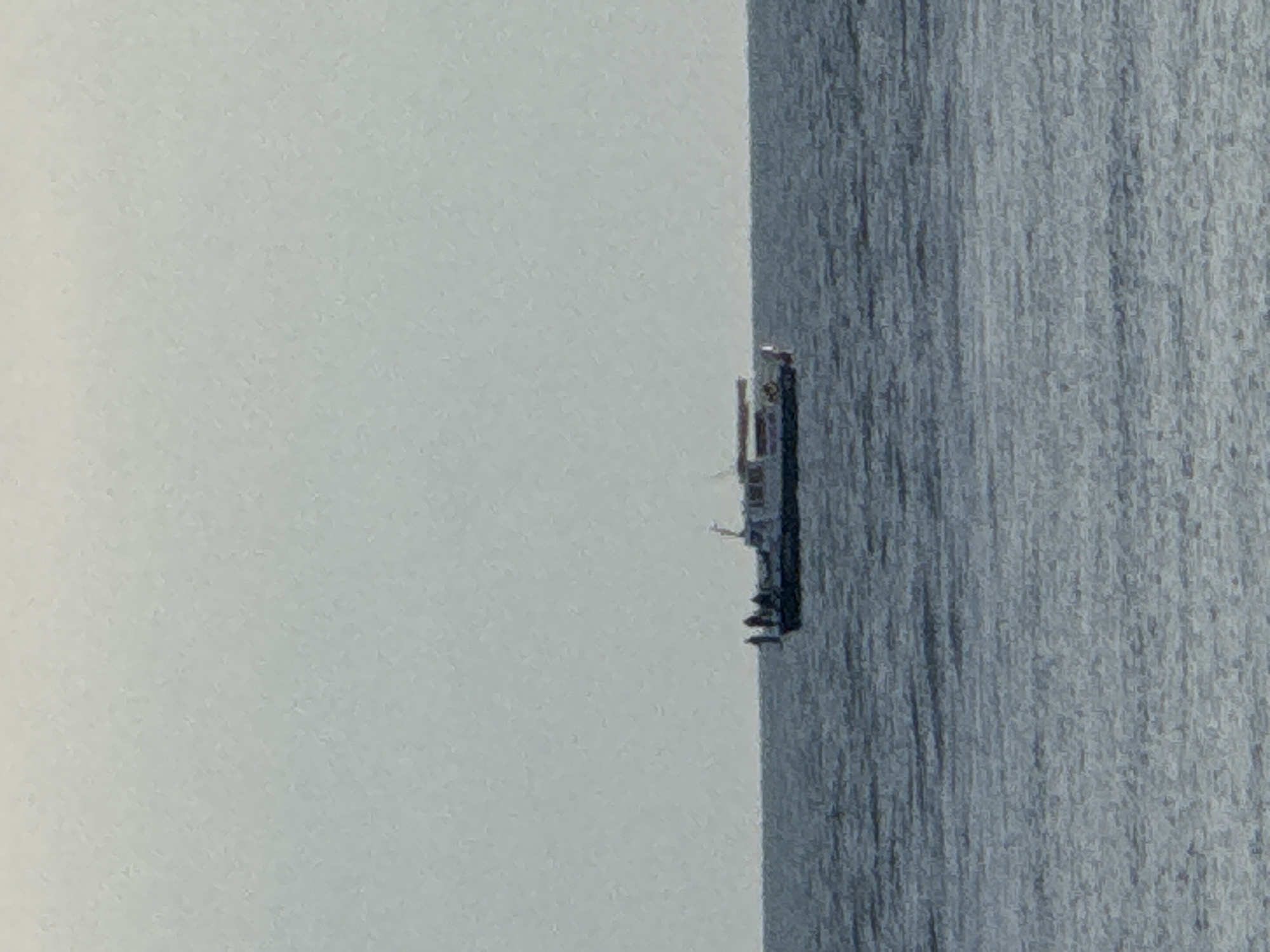 A distant shot of a ship is positioned between a body of water and a white, overcast sky. The ship is dark and vertically oriented within the frame. The water occupies the right side of the image and is textured with linear patterns, possibly waves or reflections. The white sky takes up the left side of the image, with no discernible features.