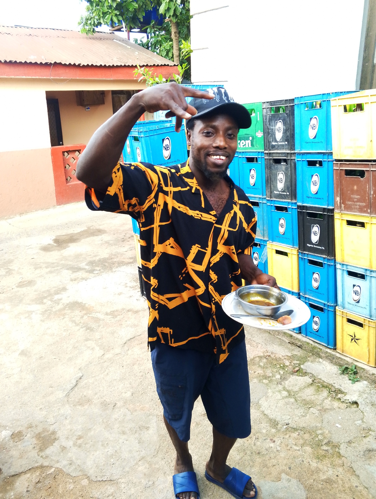 A man stands outdoors, smiling and gesturing with his right hand while holding a plate of food in his left. He wears a black cap, a black shirt with a yellow pattern, blue shorts, and blue sandals. He holds a white plate with a metal bowl containing liquid, along with a spoon and a piece of food on the side. Behind him is a stack of colorful crates, bearing the logo "NB." The background shows a building with a red corrugated metal roof and a white wall with red trim. The location is likely in Jos, Nigeria, given the context of the crates and the person's appearance. The ground is concrete.