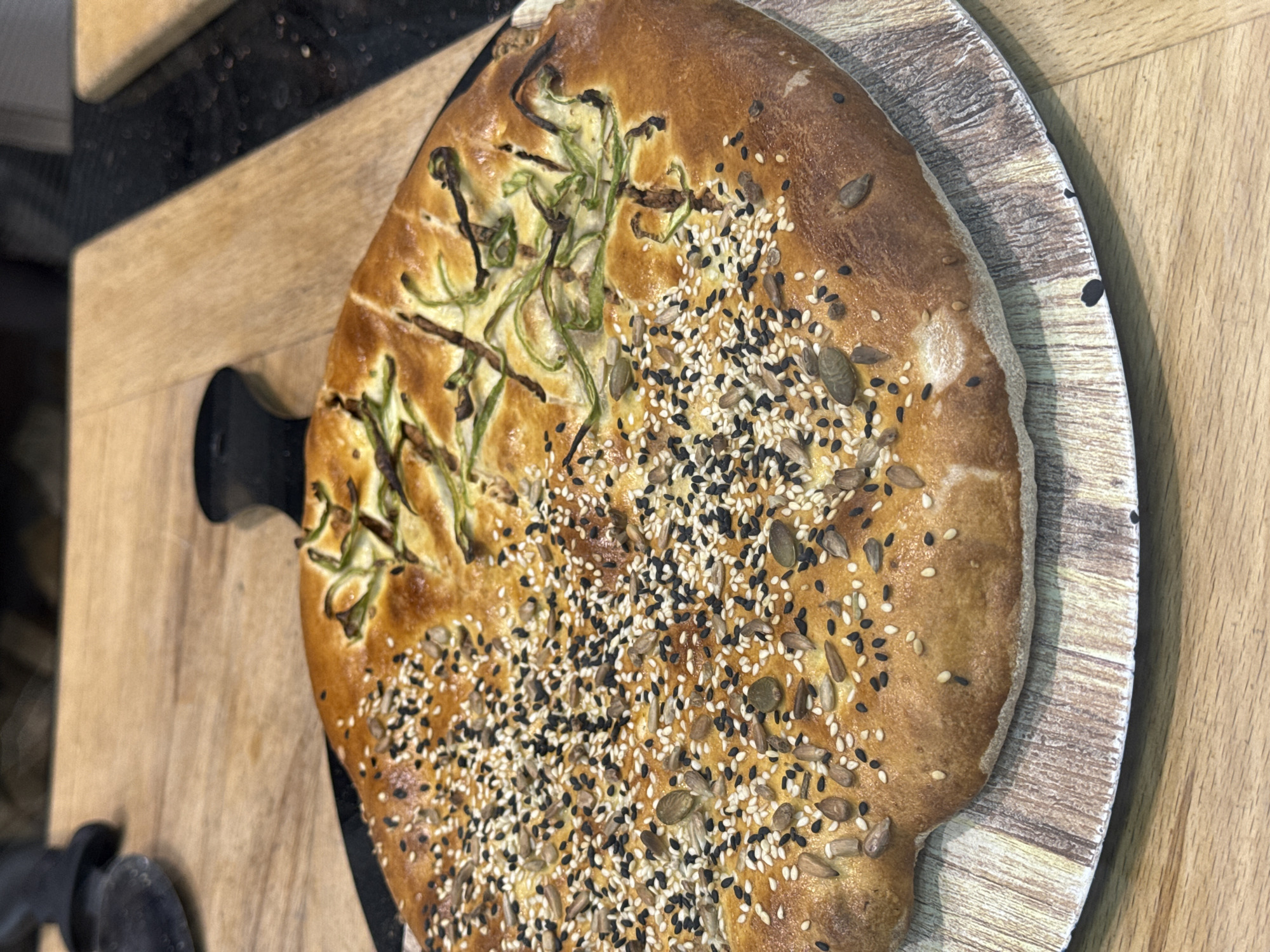 A close-up, medium-shot image shows a round loaf of bread, predominantly golden-brown with various toppings, sitting on a circular, wooden serving board with a black handle, which rests on a rectangular wooden surface. The bread is topped with what appears to be long, thin strands of green vegetables on one side, resembling peppers, and a mixture of black, white, and brown seeds (such as sesame, sunflower) on the other side. The bread's surface has a slightly irregular texture and appears baked. The wooden surfaces are a light-brown color with visible grain. The background shows a glimpse of a black countertop. The image appears to be taken in an indoor setting. There are no people visible. The location of Aparan, Armenia is not evident from the image.