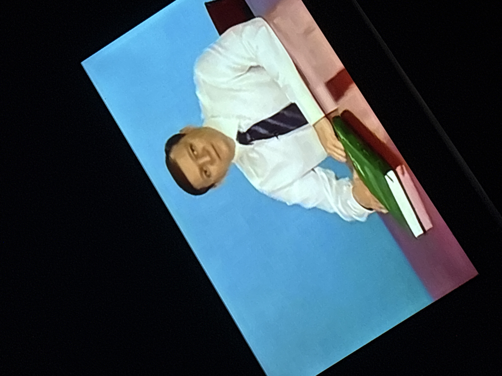 The image shows a man in a white dress shirt and a dark tie sitting at a desk. He is looking to his left and has his left hand resting on a green object, possibly a book or folder. The desk is covered in a pinkish-red surface, and behind the man is a bright blue background. The scene is viewed from an angle, with a black border around the image. It is not possible to determine the location in the city of Dnipro, Ukraine from the provided image.