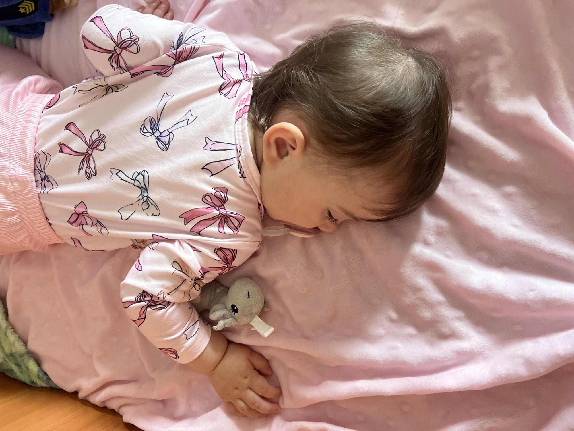 A baby is lying on a pink blanket, wearing pink pajamas with a bow pattern. The baby has dark hair and is sleeping with a pacifier in their mouth and a gray stuffed animal next to their hand. The image does not contain any information to place the scene in Elena, Bulgaria.