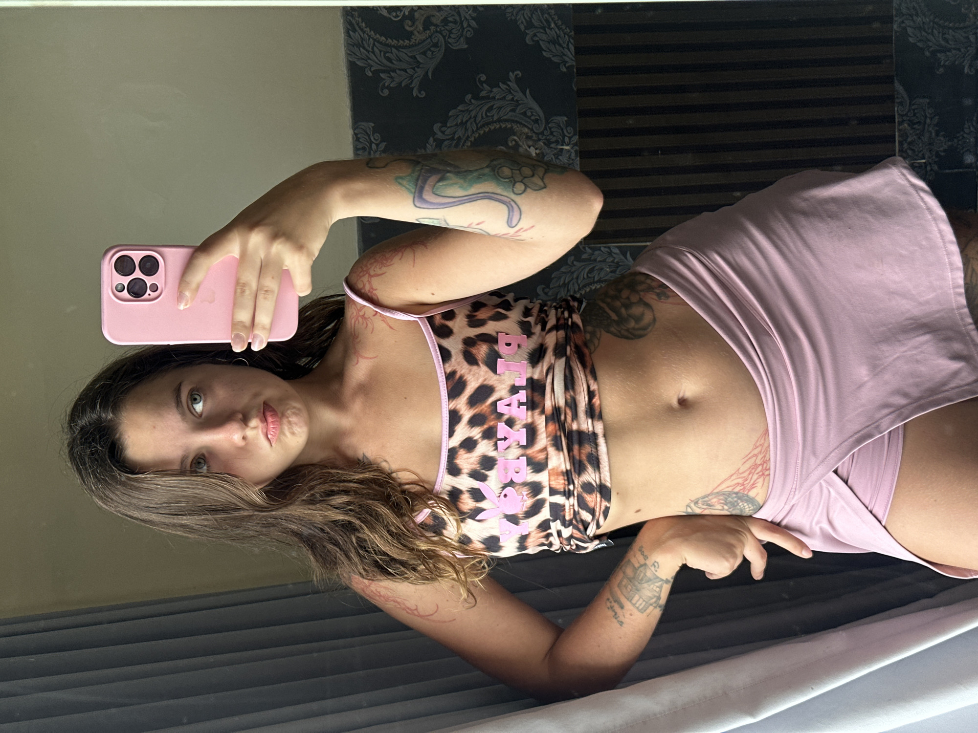 A young woman with brown hair and blue eyes is taking a mirror selfie. She is wearing a leopard print crop top with pink trim and a pink skirt that appears to be a skort. She has several tattoos on her arms, chest, and leg. She is holding a pink iPhone in her right hand and is looking at the camera with a pouty expression. The background is a bathroom with a patterned wallpaper and vertical blinds.