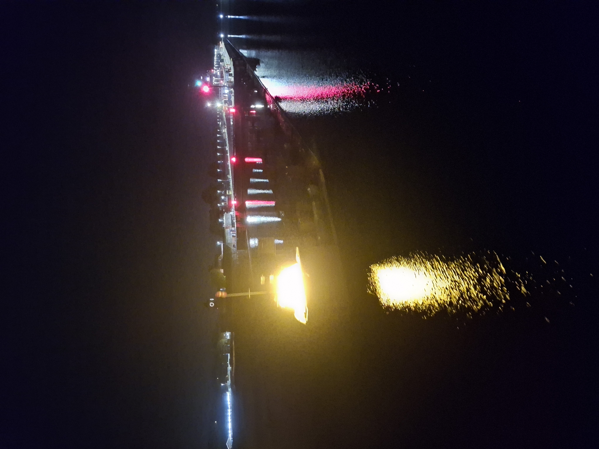 Here is a description of the image:
The image depicts a nighttime scene of a bridge extending over water. Lights line the bridge, with reflections visible in the water below. Red lights are also present on or near the bridge. A bright, yellow light is prominent in the foreground, casting a strong reflection on the water's surface. The surrounding environment is dark, with limited visibility beyond the bridge and its immediate vicinity. The city and state are unknown.