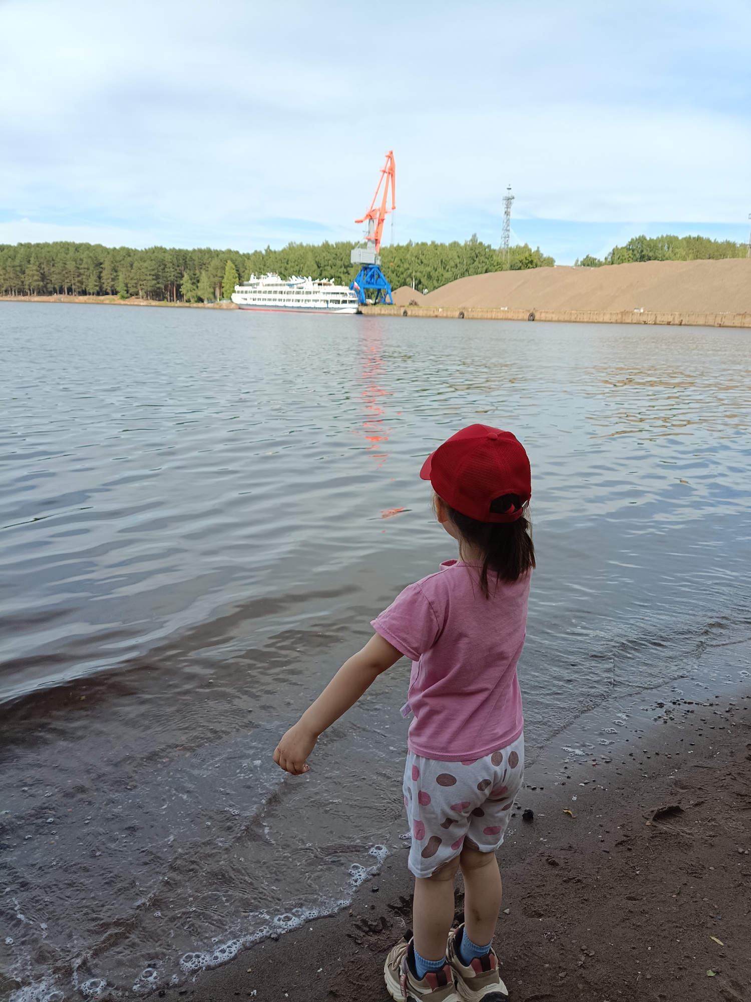 A young child wearing a red baseball cap, a pink t-shirt, and patterned shorts stands at the edge of a body of water, with their right hand extended towards the water. The child is facing away from the camera, looking out at the water and the industrial scene beyond. In the background, a large orange crane stands near a white passenger ship docked on the opposite shore. Beyond the ship and crane, a dense forest lines the horizon, with a large pile of sand to the right of the crane. The sky above is a light blue with scattered clouds. The foreground shows a sandy and pebbly shoreline with small waves lapping at the edge. The image appears to be taken in the port area of Chaikovsky, Russia, given the industrial elements and the overall setting.