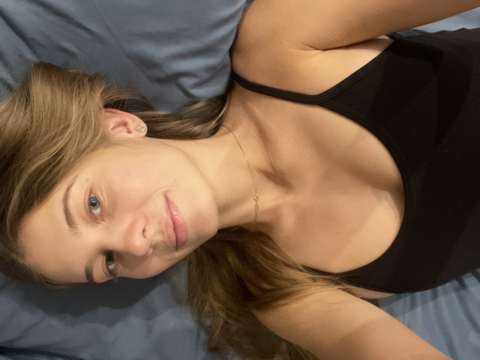 A young woman reclines on a blue surface. She has light brown hair, pale skin, and blue eyes. She is wearing a black tank top and a delicate gold necklace. Her face is turned slightly towards the camera, displaying a faint smile. An earring is visible in her left ear. The soft lighting suggests a relaxed, intimate setting.