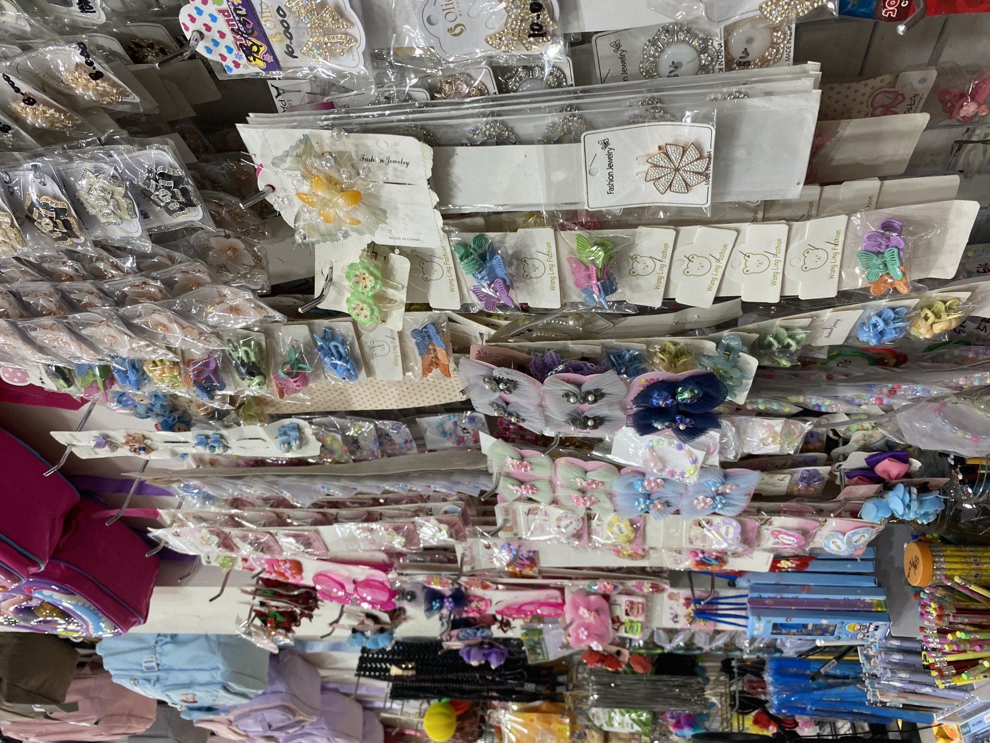 The image depicts a brightly lit, cluttered stall at a market, likely in Lamteumen Barat, Indonesia. The stall is filled with a diverse array of small, colorful accessories, primarily hair clips, jewelry, and possibly stationery. The items are predominantly displayed on hanging racks, some individually packaged in clear plastic, others in bulk.
Dominating the upper portion of the image are several children's backpacks, one vibrant pink and another a light blue, suggesting a focus on younger customers. Below these, a dense collection of hair accessories is visible, including butterfly-shaped clips, flower-shaped barrettes, and small, fuzzy hair ties. The colors range from pastel pinks and blues to bright greens and oranges.
Further down, in the lower part of the frame, a collection of pens and pencils is neatly arranged in containers, hinting at a stationery section. There are also various pieces of costume jewelry, such as brooches and earrings, adorned with rhinestones and pearls. The overall impression is one of abundance and variety, characteristic of a busy local market. The background appears to be a simple wall, with overhead lighting illuminating the merchandise. There are no people clearly visible in the frame.