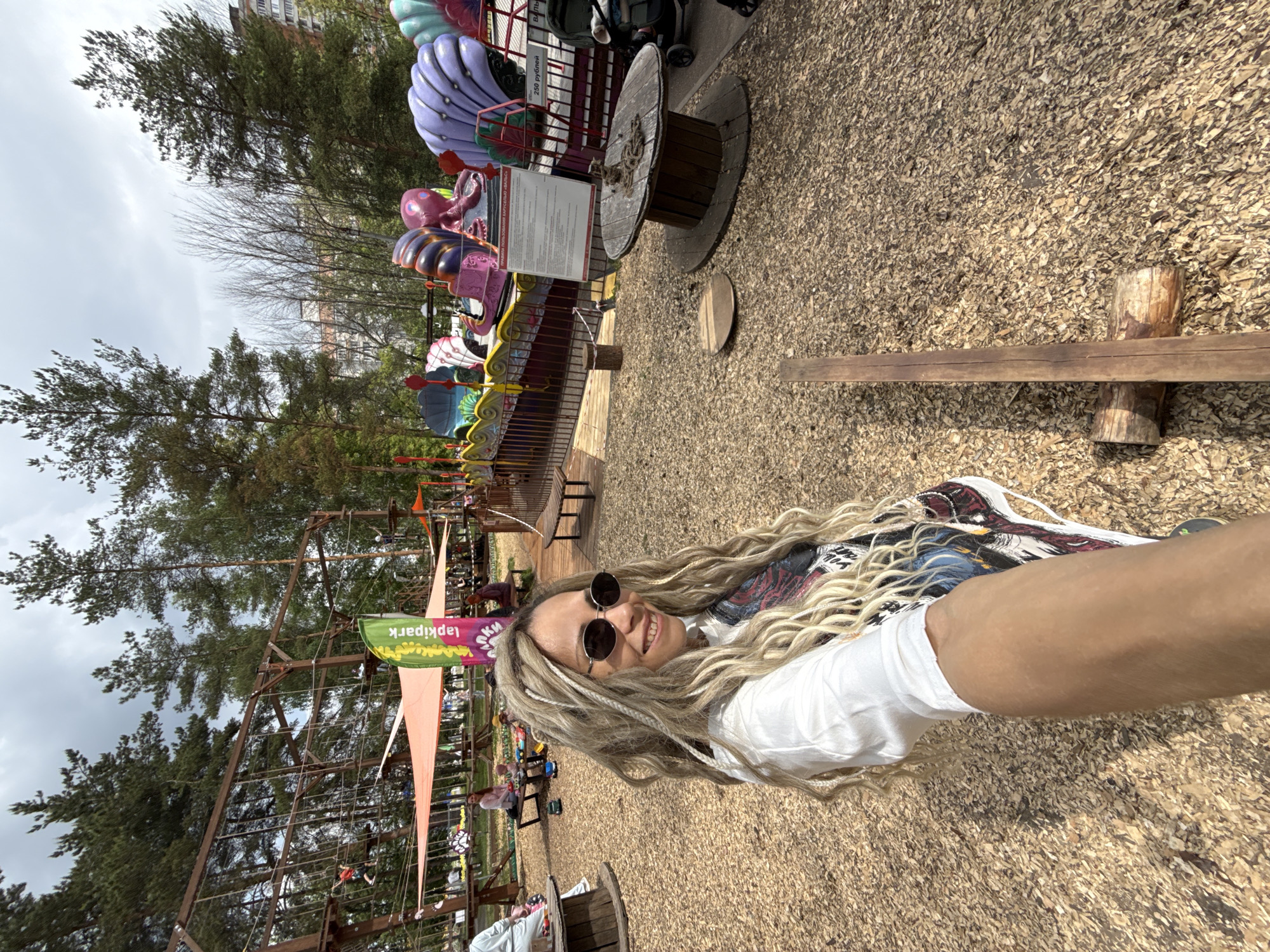 A selfie-style image captures a smiling woman with long, light-colored dreadlocks, wearing dark sunglasses and a white t-shirt, at an outdoor amusement or adventure park. The woman, positioned prominently, faces the camera with a joyful expression.
The park setting features several attractions, including colorful rides with seashell and octopus designs, and a wooden adventure course structure with ropes and nets. The ground is covered with wood chips. In the background, lush green trees are visible, along with distant multi-story residential buildings under a cloudy, overcast sky.
The scene suggests a casual visit to an outdoor recreational area. Notable details include a large wooden spool, wooden benches, and other visitors enjoying the park in the distance.
Visible text includes a green and purple banner displaying "Лапкипарк" and "lapkipark". Another white sign near one of the rides shows "250 рублей" (250 rubles), indicating a price. A larger white sign next to it also contains Russian text, likely detailing information or rules for the attraction. The location is contextualized as Шебунино, Russia.