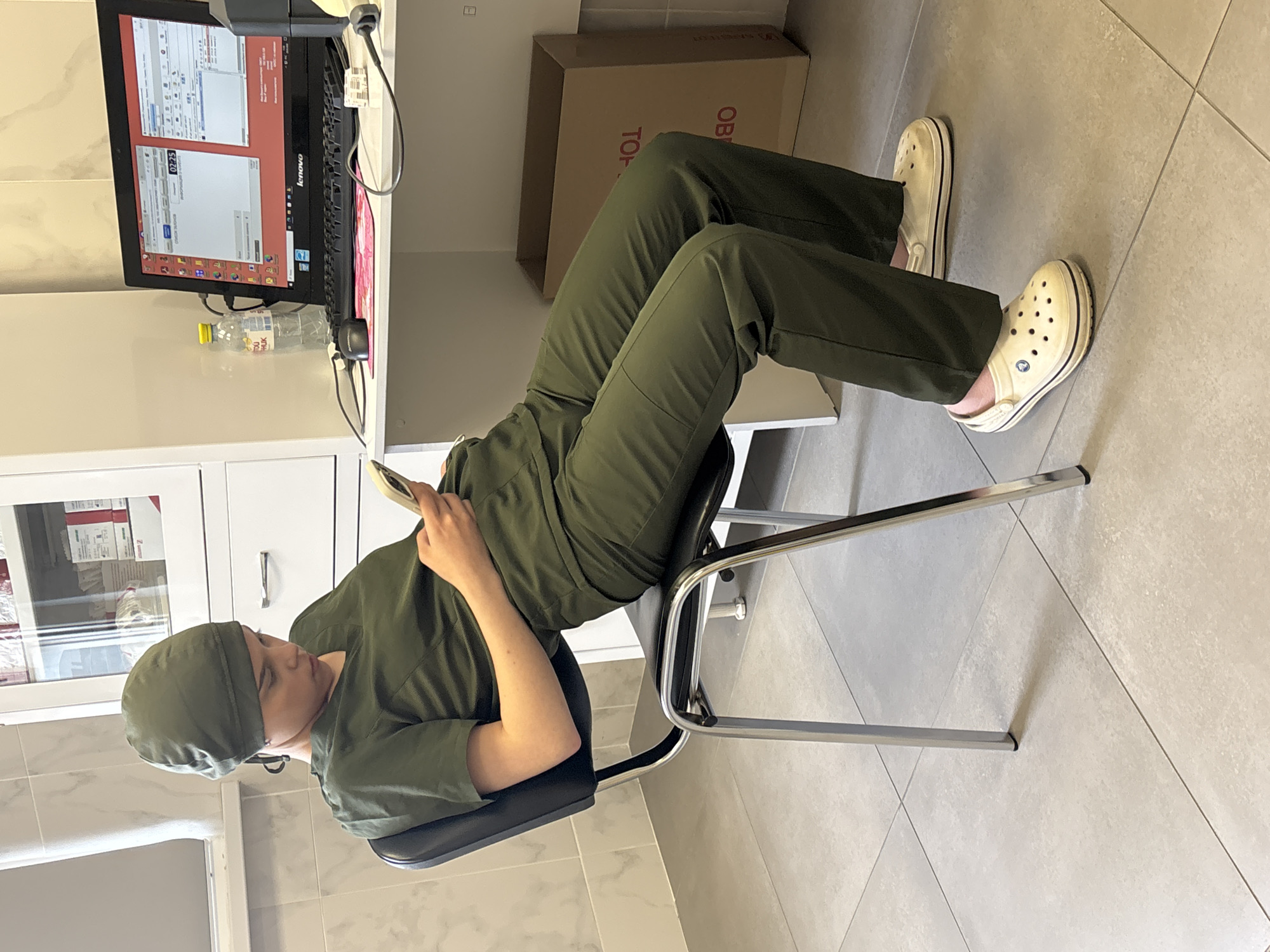 A person wearing green scrubs and a scrub cap sits in a metal-framed chair with a black seat in what appears to be a medical or clinical setting. The person is looking at a smartphone in their hand. They are wearing light-colored Crocs.
Behind the person is a computer monitor displaying software. A bottle of water is visible next to the monitor. There are white cabinets to the left of the person, and a cardboard box is to their right. The floor is tiled. The specific location within Saint Petersburg, Russia is not discernible from the image.