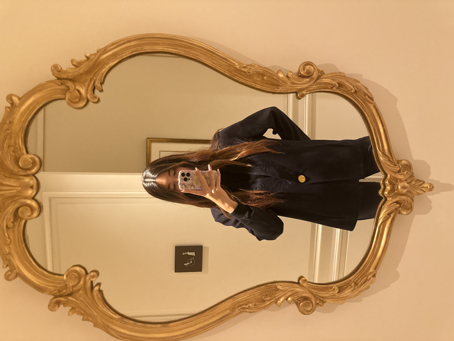 A person is seen in a mirror taking a selfie. The mirror has a decorative, ornate gold frame. The person has long, dark hair and is wearing a dark jacket with a gold button. Behind the person in the reflection, a framed picture and a door can be seen. A small, black sign featuring a white figure and wheelchair symbol is mounted on the wall near the bottom of the mirror. The wall is a light beige color. The image is rotated clockwise.