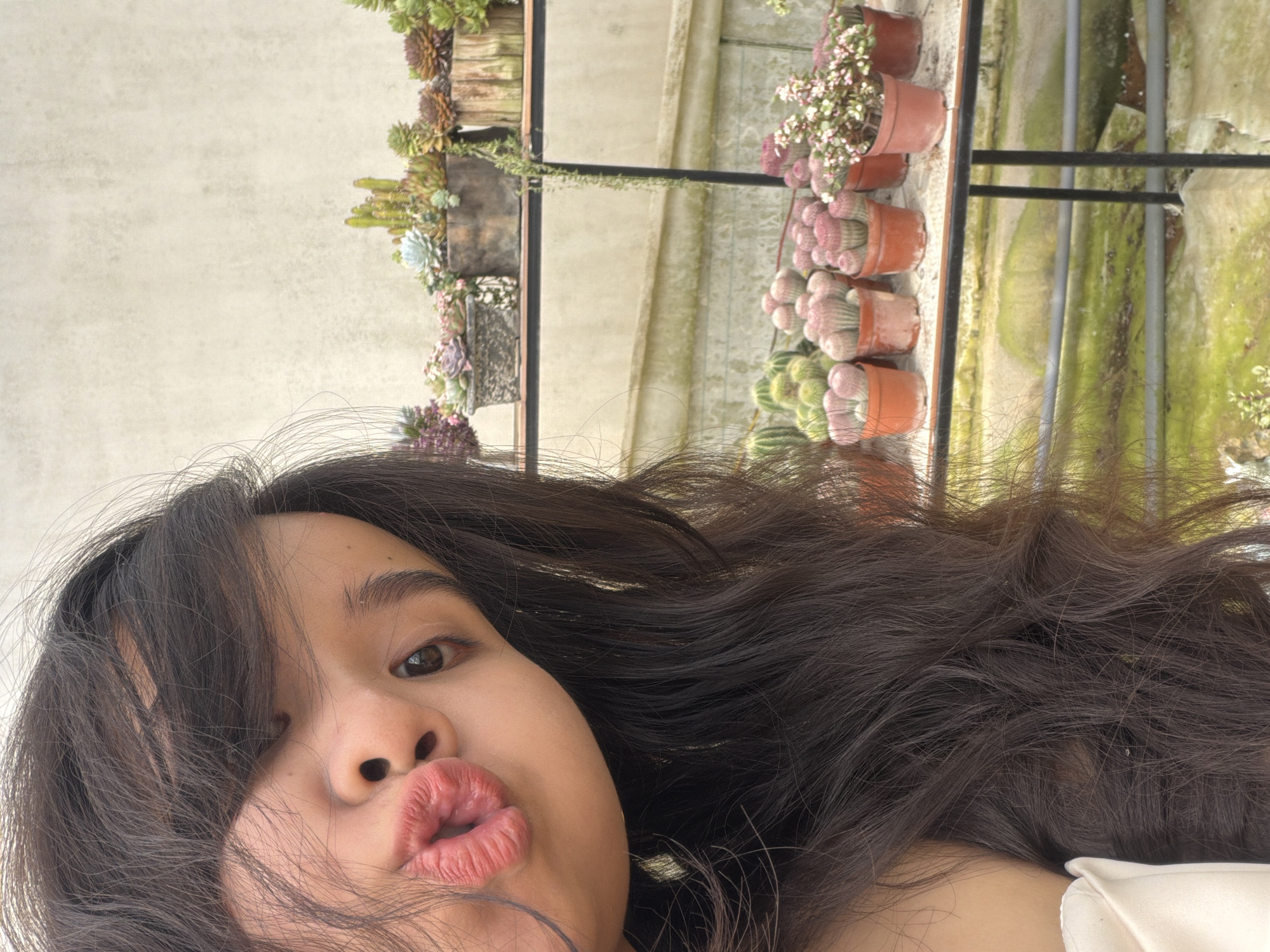 A close-up, selfie-style portrait shows a young person with dark, flowing hair puckering their lips. The person has light brown skin, dark eyes, and full lips. The background, slightly blurred, reveals a collection of potted plants on tiered shelves, including cacti and succulents. The scene likely takes place in a greenhouse or outdoor garden setting. The specific location of Măng Đen, Vietnam, is not evident in the image itself.