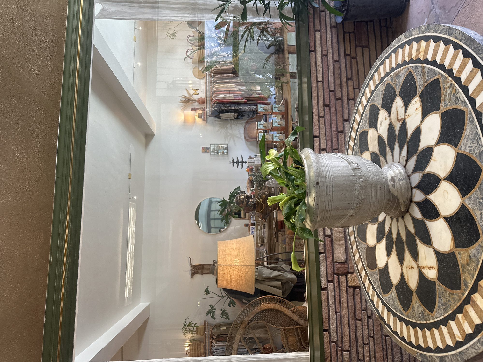 This image shows the interior of a store, captured through a mirror or glass pane. The store features a white interior with displays of various items, including textiles, decorative objects, and plants. A lamp with a tan lampshade is visible, along with a wicker chair. The reflection gives the impression of a bright and airy space. In the foreground, next to what appears to be a brick wall, is a large, circular table with a geometric black and white pattern. On the table sits a gray urn-like vase containing a green plant. The image provides a glimpse into the design and aesthetic of the store, which appears to be in a city of Unknown, Unknown.
