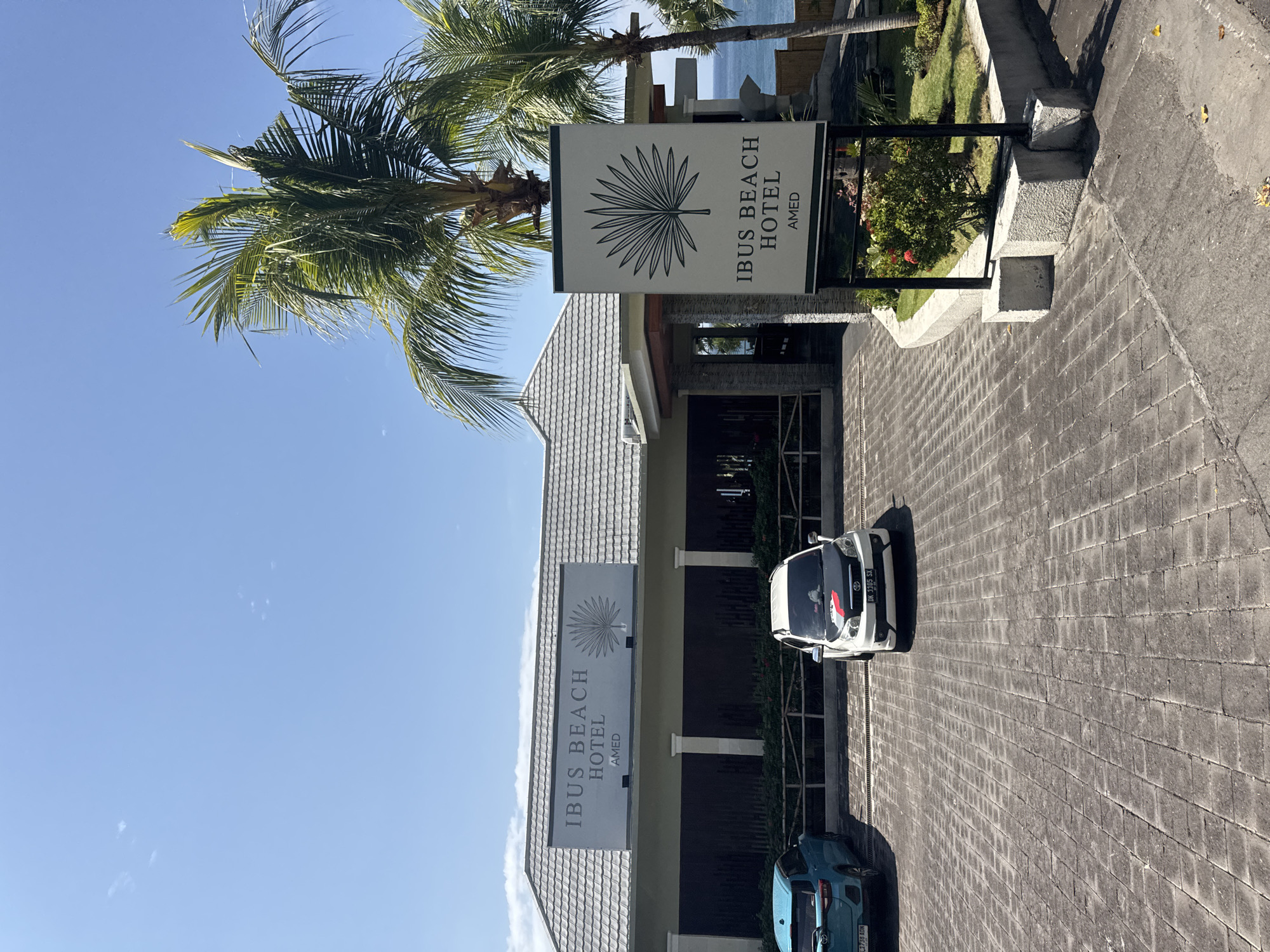 The image depicts the entrance of the Ibus Beach Hotel in Abang, Indonesia. The hotel has a modern architectural style with a gray tiled roof and a light-colored facade. A prominent sign with the hotel's name and logo, a stylized palm frond, is displayed on the front of the building. Another similar sign is placed on a stand in front of the hotel, beside a palm tree.
In the foreground, a paved driveway with interlocking square stones leads to the hotel entrance. Two cars are parked on the driveway: a blue hatchback on the left and a white SUV with a black hood on the right. The white SUV has a license plate that reads "DK 3305 SX".
The environment is sunny with a clear blue sky. Palm trees are visible, suggesting a tropical location. In the background, a glimpse of the ocean can be seen, indicating the hotel's proximity to the beach. The scene is clean and well-maintained, with some greenery and landscaping around the hotel entrance. There are no people visible in the image.