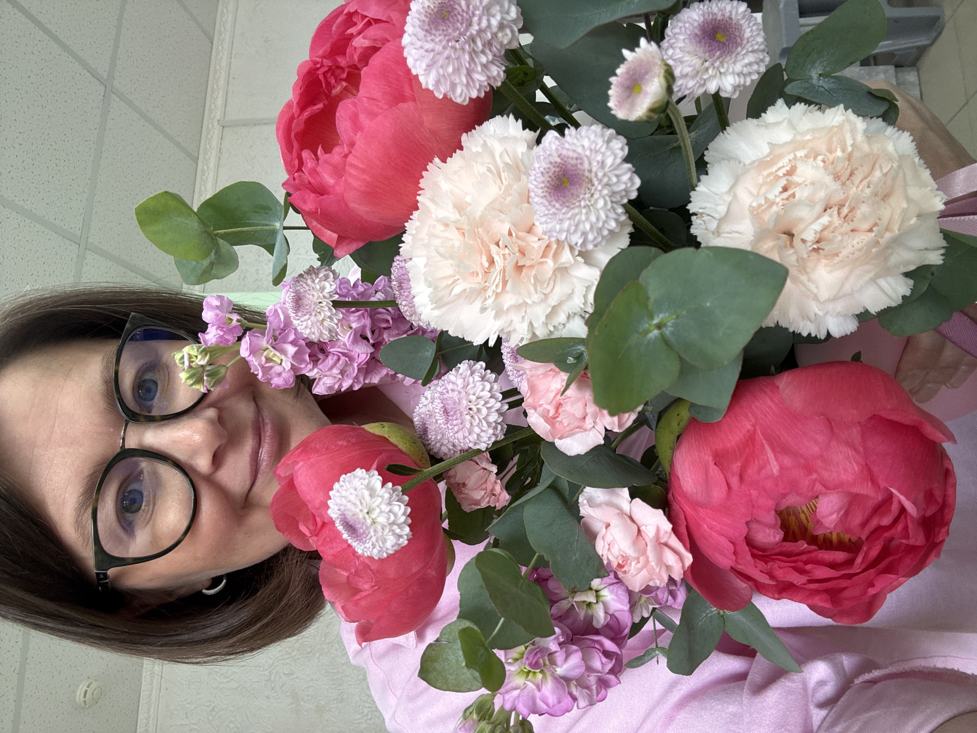 A person, likely female, wearing black-rimmed glasses, is holding a bouquet of flowers. The flowers consist of pink peonies, light pink carnations, small lavender-colored pompom chrysanthemums, and eucalyptus leaves. The person is wearing a pink shirt. The background appears to be an interior with a neutral-colored wall. There is no indication in the image that the location is Izhevsk, Russia.