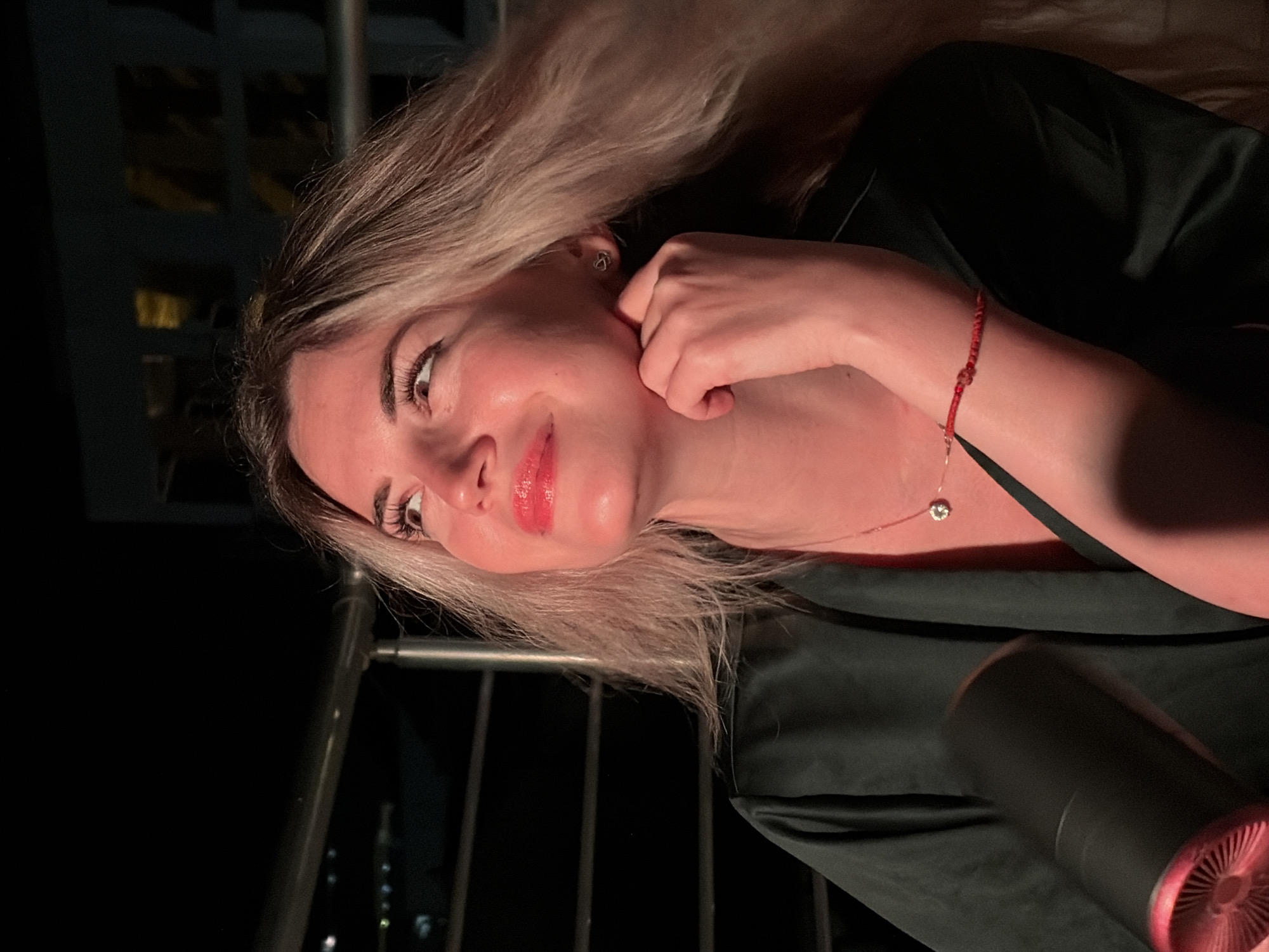 A young woman with blonde hair and red lipstick is captured in a low-light, outdoor setting. She is wearing a dark green satin robe and a delicate necklace with a small diamond pendant. A thin red string bracelet is visible on her wrist. Her right hand is gently touching her chin. In the foreground, a black cylindrical object with a red, textured end is held by her left hand, its function unclear due to the low light and motion blur. The background is dark, with hints of railings and distant lights suggesting an urban environment, possibly a balcony or rooftop overlooking Budva, Montenegro. The lighting is warm and dramatic, casting a reddish hue on her skin.