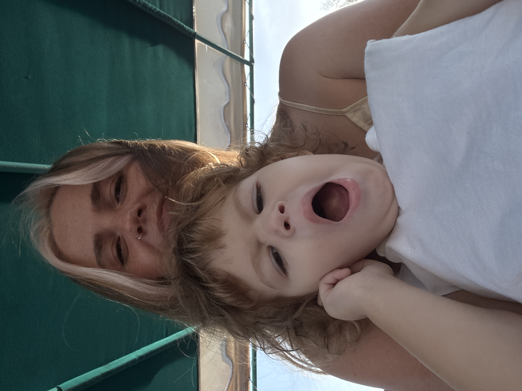 A selfie taken from a low angle shows a woman and a young child. The woman, in the background, has blonde hair with some lighter streaks and is looking at the camera with a slight smile. She is wearing a light-colored camisole. The child, in the foreground, is leaning against the woman and appears to be yawning. The child has curly brown hair and is wearing a white t-shirt. They are both under a teal canopy, suggesting they are outdoors, possibly in a shaded area like a park or a covered seating area. The lighting suggests it is daytime. There is no visible indication of the location in Choeng Thale, Thailand.