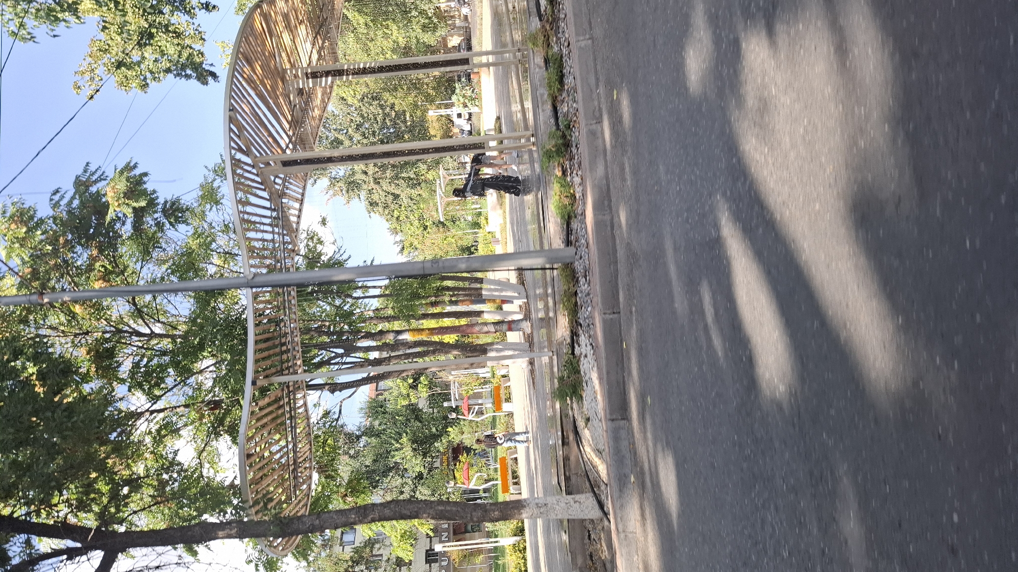 The image depicts a street scene in Shymkent, Kazakhstan. In the foreground, a paved road shows dappled sunlight, suggesting the presence of trees overhead. A concrete curb separates the road from a walkway. On the walkway, there is a modern, open-air structure with a curved, slatted roof supported by metal poles. Several trees are planted along the walkway, some with white-painted lower trunks.
Further back, behind the modern structure, there are benches and what appear to be colorful, abstract sculptures or play equipment. A person in dark clothing, possibly a woman wearing a headscarf and long dress, is standing under the modern structure, interacting with another person who is mostly out of frame. A third individual, wearing jeans and a dark top, stands near some benches in the mid-ground. In the background, buildings and more trees are visible, indicating an urban environment. The overall impression is of a public space, possibly a park or a rest area along a street.