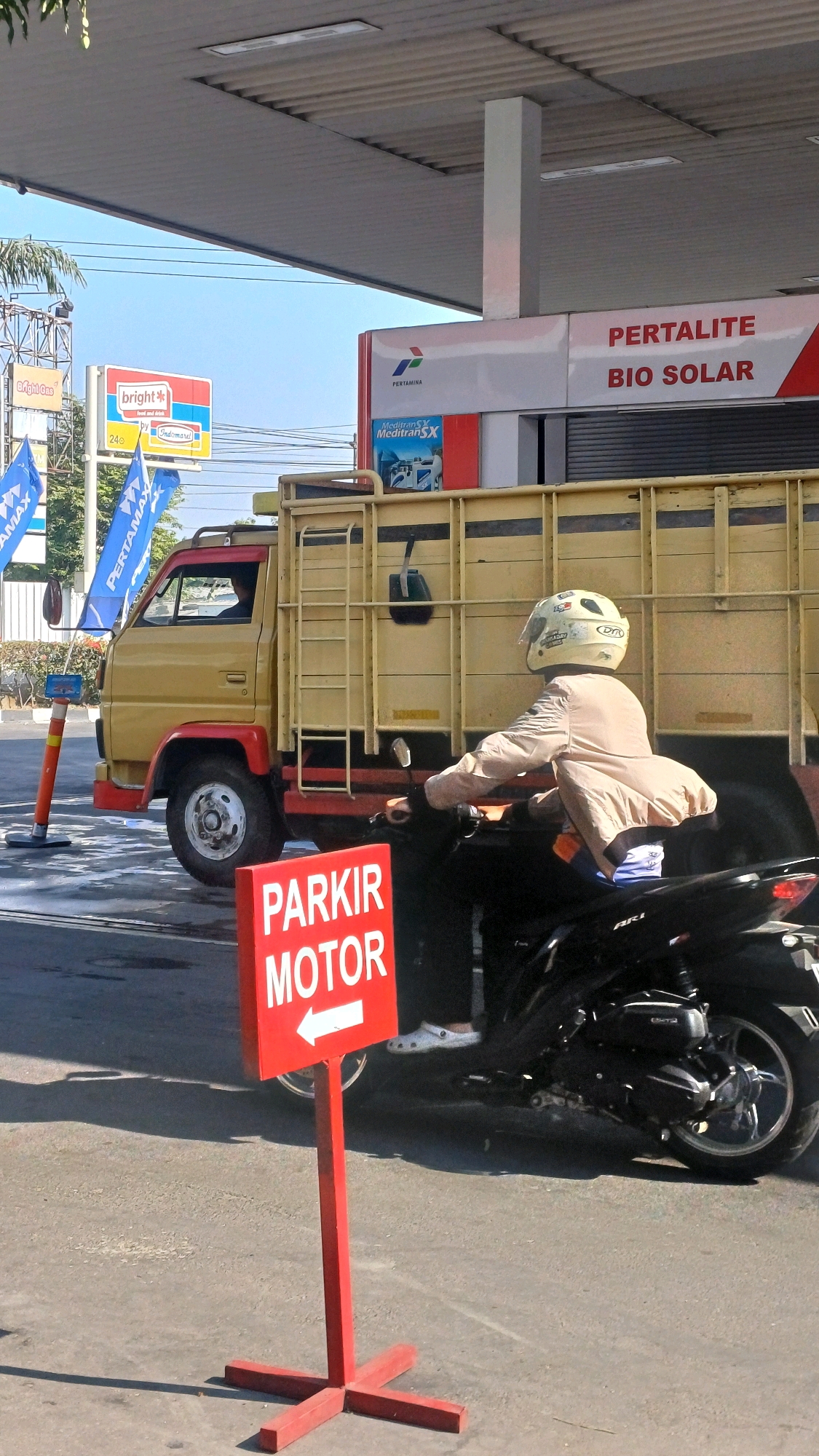 At a Pertamina gas station in Pabelan, Indonesia, a person on a motorcycle wearing a beige jacket and a white helmet is parked. The motorcycle is black and parked in front of a red sign that reads "PARKIR MOTOR" with a white arrow pointing left. Behind the motorcycle is a yellow truck with a red frame around the cab. The truck has a ladder on its side and a black bag hanging from it. The gas station canopy is visible above, with signs for "PERTALITE" and "BIO SOLAR." In the background, there are blue flags with "PERTAMAX" written on them and a "Bright Gas" sign. The ground is wet, suggesting recent rain or cleaning. The sky is clear and blue.