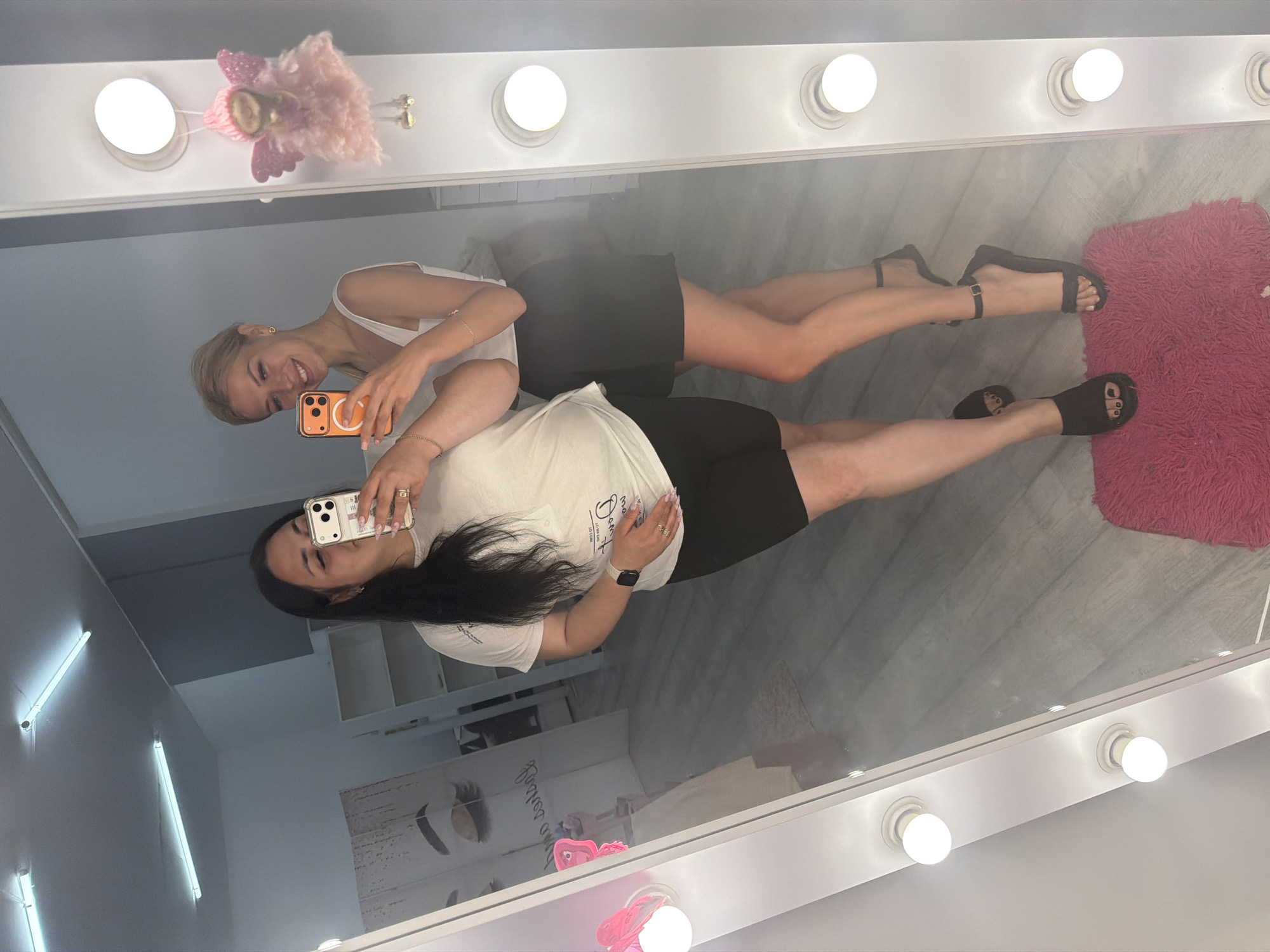 Two women are taking a mirror selfie in what appears to be a beauty salon or studio in Kyiv, Ukraine. The woman on the left has long dark hair and is wearing a white t-shirt and black shorts. She is holding a smartphone in her left hand, taking the photo. The woman on the right has blonde hair and is wearing a white tank top and a black skirt. She is also holding a smartphone, and smiling at the camera. Both women are wearing sandals. The mirror is framed by a series of lights. In the background, there are shelves and a wall with a graphic of an eye and eyelashes. A pink fluffy rug is visible on the floor in front of the mirror.