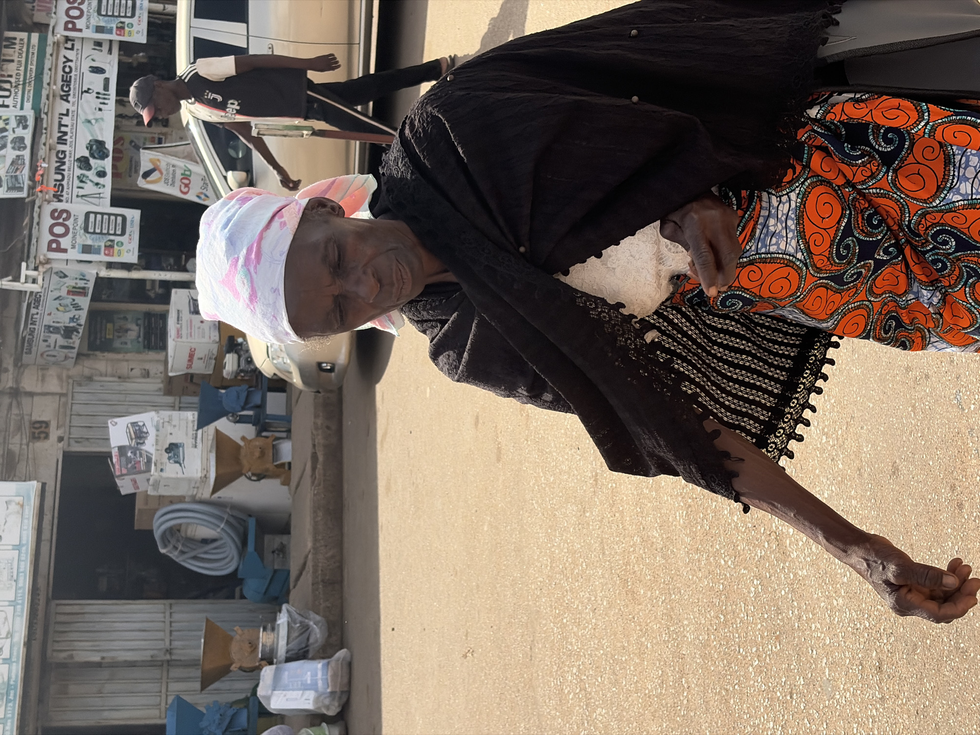 This media file captures an outdoor scene in Jos, Nigeria. The primary subject is an elderly woman wearing a black shawl and a colorful, patterned skirt, with a pink and white headscarf. She is looking towards the camera with her eyes closed, appearing relaxed or possibly sleeping. Her left arm is extended, resting on the ground.
In the background, a man wearing a black and white shirt and a red baseball cap is walking away from the camera. To the left of the scene, there are various commercial displays and signage, including "SAMSUNG INT'L AGECY", "FUJIFILM AUTHORISED FUJI DEALER", and "POS". These indicate a commercial area with shops selling electronics and related services. Further down the street, boxes and machinery, possibly agricultural equipment like grinders, are visible, suggesting a marketplace or a business that sells such items.
The time of day appears to be late afternoon or early evening, as indicated by the long shadows cast on the ground and the warm, golden light. The weather seems to be pleasant, possibly dry. The overall impression is that of a bustling street in a Nigerian city, with everyday life unfolding. The presence of the elderly woman suggests a moment of rest or quiet amidst the activity.