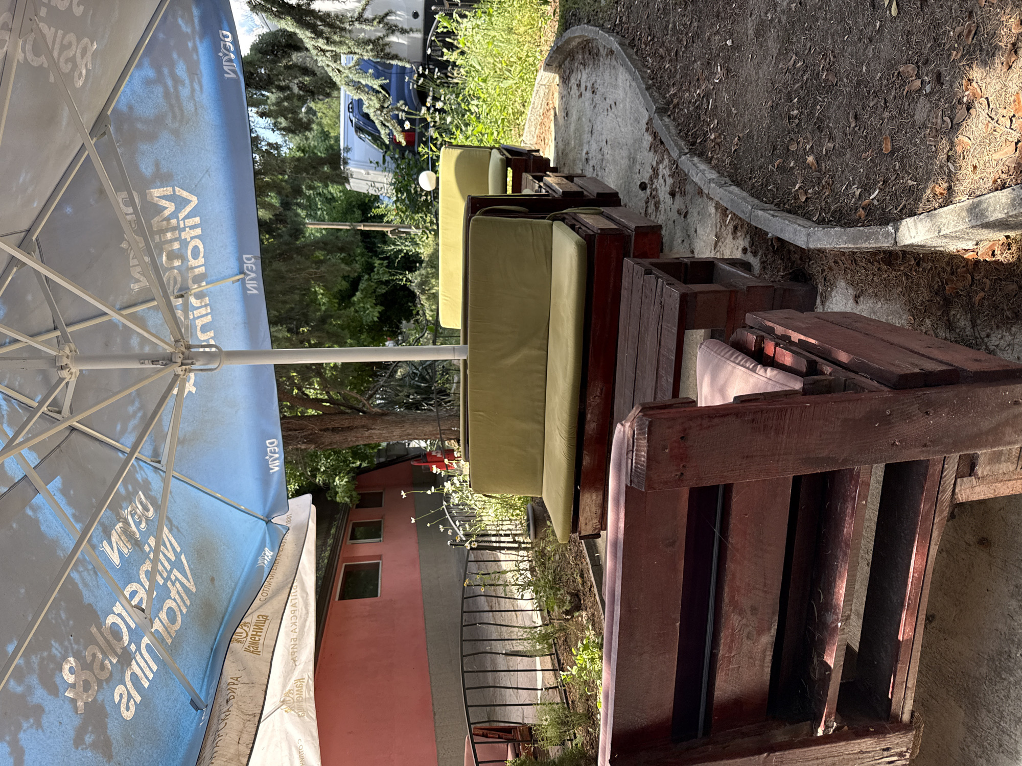 The image depicts an outdoor seating area, likely at a cafe or restaurant, with wooden benches and tables under a large blue umbrella. The umbrella has white text printed on it, appearing to be a brand name or advertisement, with some sections reversed due to being viewed from underneath. The seating area is surrounded by a small garden with green plants and white flowers, and a black metal fence. In the background, a pink building is visible, along with trees and a parked white van and a dark blue vehicle. The ground is a mix of concrete and dirt, with fallen leaves scattered around. The overall impression is one of a casual outdoor space in a temperate climate. The specific location is identified as Varna, Bulgaria.